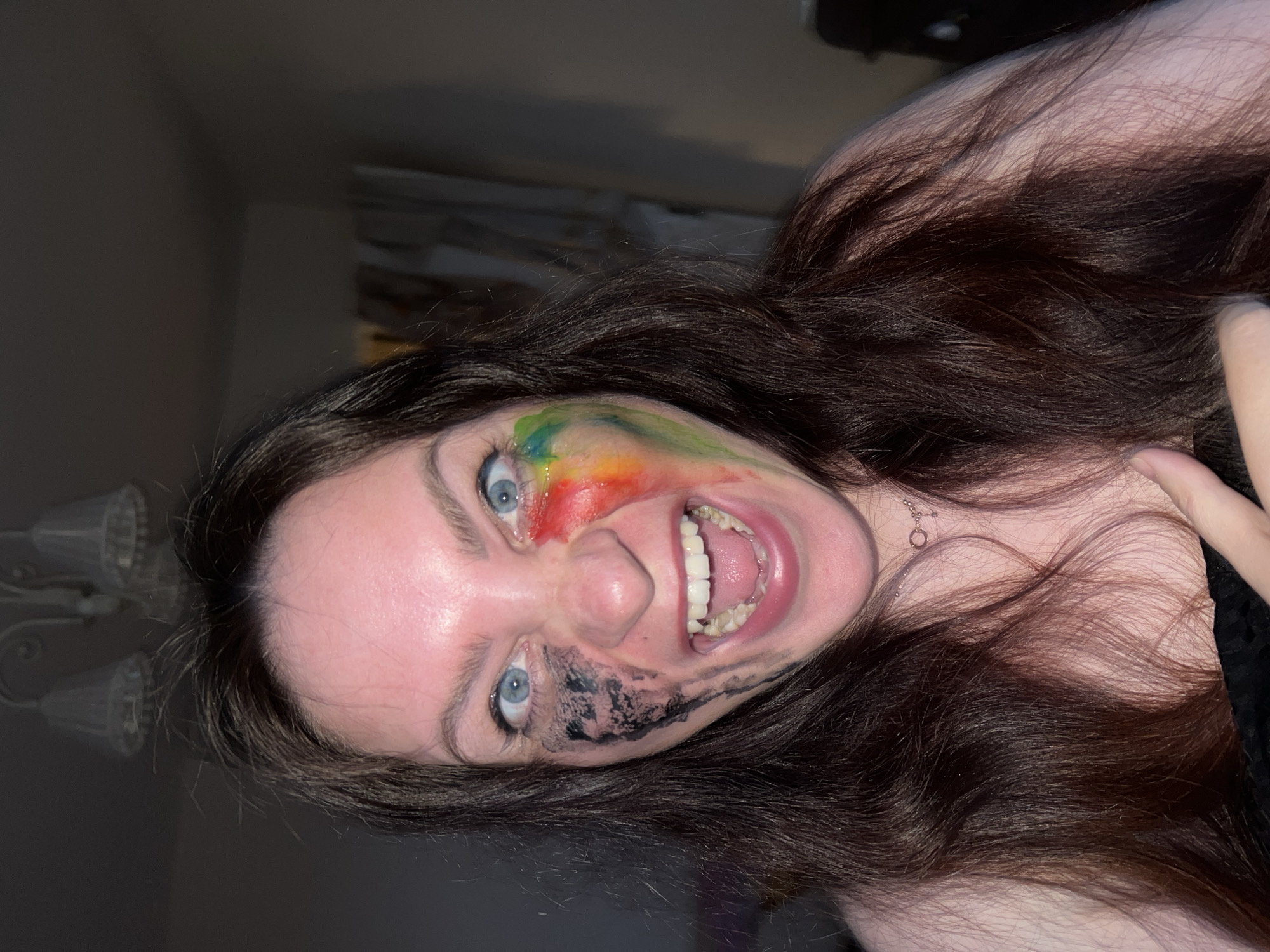 A young woman with long, dark hair and blue eyes is pictured in a close-up shot. Her face is painted with colorful streaks of paint on one side and black smears on the other. She is grinning widely, revealing her teeth. The background is blurry, suggesting an indoor location. The lighting is dim. There is no indication this photo was taken in Noviy, Russia.