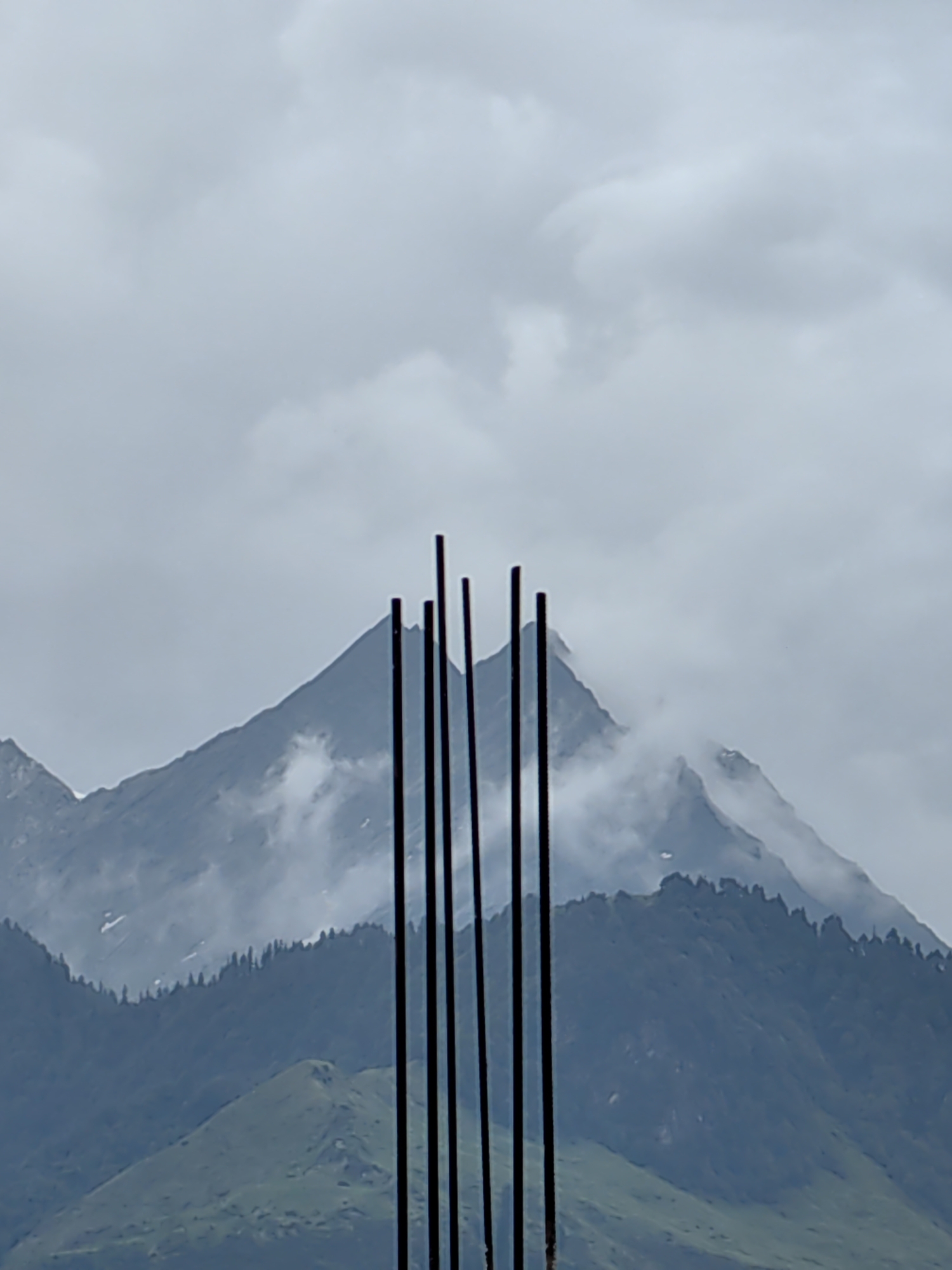 The image shows a mountainous landscape with a cloudy sky. In the foreground, there are several vertical black poles, possibly part of a construction site. The mountains in the background are covered in mist and fog, with some patches of green vegetation visible on their lower slopes. The overall mood is somber and atmospheric, suggestive of a damp or overcast day in a high-altitude region. The location is Vashisht, India, known for its hot springs and scenic mountain views.
