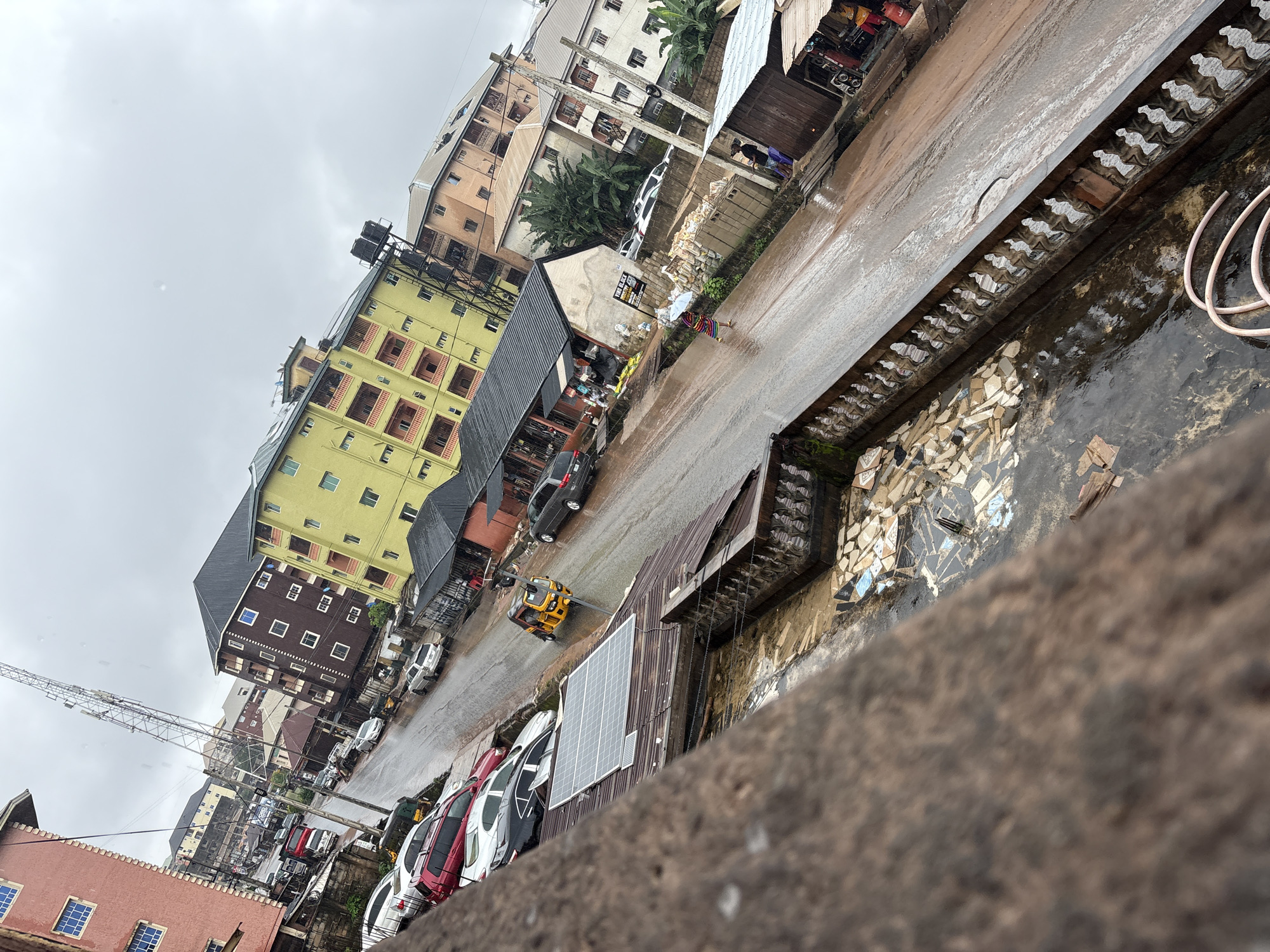 The image shows a street scene in Nkpor, Nigeria, under a cloudy sky, likely after rain given the wet pavement. The street is lined with multi-story buildings of various colors, including a red, a yellow, and a brown one. Several cars are parked along the side of the road, and a yellow three-wheeled auto-rickshaw is in motion. Some of the buildings appear to be residential, with balconies and windows. One building has solar panels on its roof. There are also smaller market stalls or shops along the street, some with awnings. A few individuals are visible, including a person walking in the street and another standing near a stall. The foreground shows a weathered concrete structure with decorative railings, overlooking the street. Debris and some broken tiles are scattered on the ground near the foreground structure. A telecommunications tower is visible in the distance.