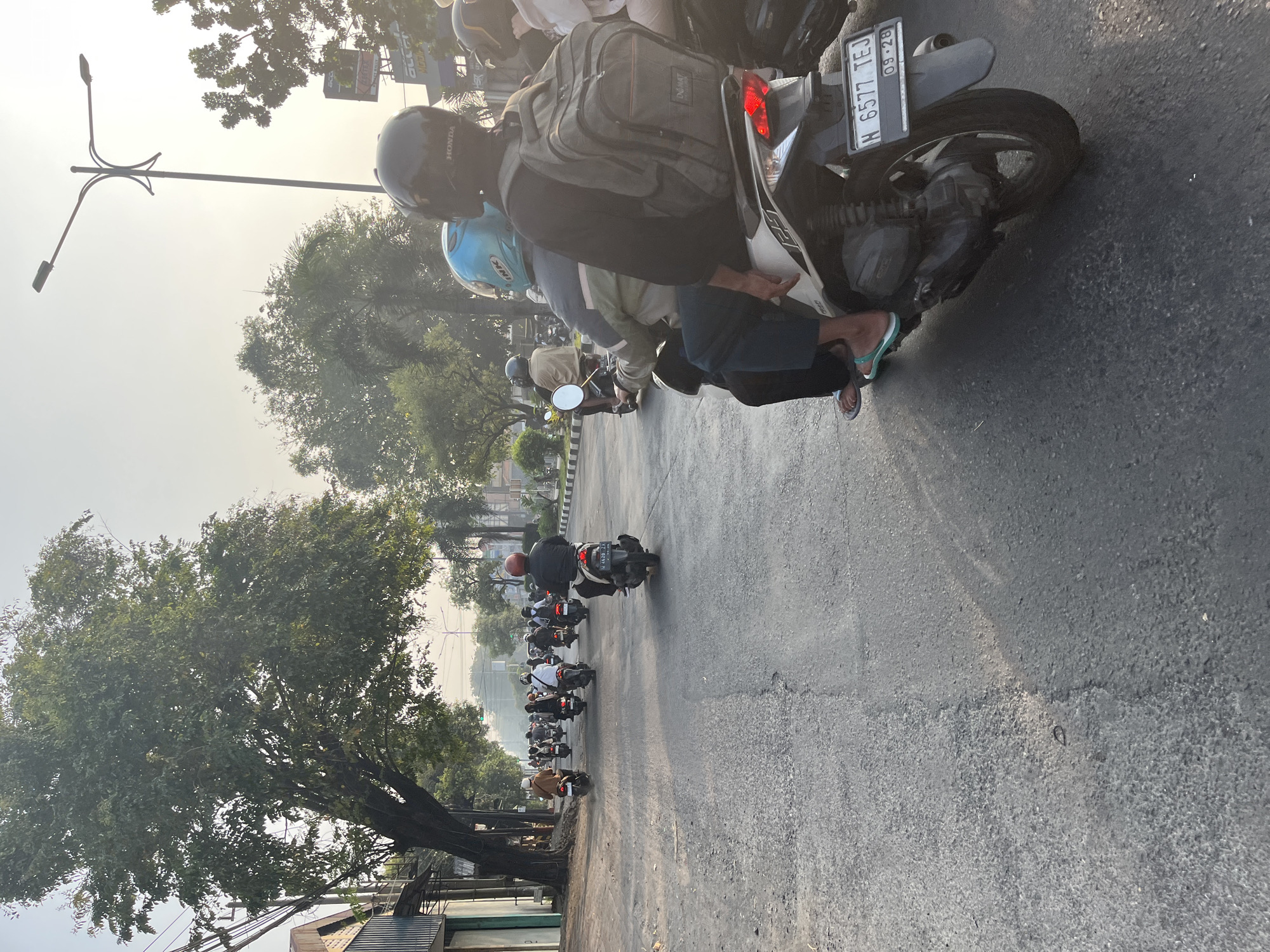 The image depicts a street scene in Tenggulunan, Indonesia, with a procession of motorcycles on a paved road. The time of day appears to be late afternoon, indicated by the soft, warm light and long shadows. Several people are riding on the motorcycles, mostly wearing helmets for safety. The foreground features a white and black motorcycle with two individuals on it, one wearing a blue helmet and the other a black helmet with a backpack. The motorcycle has a license plate that reads "N 6577 TEJ" and "09-28". The road is lined with buildings on the left and trees and streetlights on the right. The overall atmosphere is that of a busy urban environment with moderate traffic.