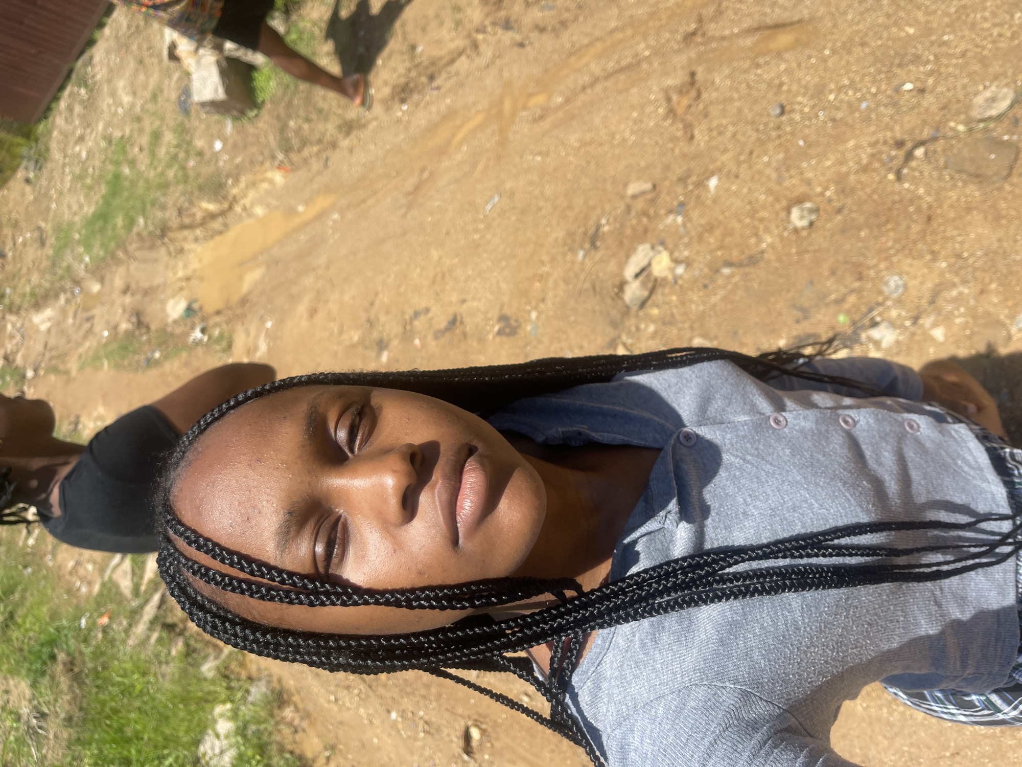 A young Black girl with braided hair is taking a selfie. She is wearing a light gray hooded top with buttons down the front, and plaid shorts. Her eyes are closed, and she has a neutral expression. Behind her, the ground is a mixture of dirt and grass, with a muddy patch in the center. Another person, wearing a black t-shirt, is visible in the background to the left, and the legs of another person in patterned shorts and sandals are visible to the right. The scene appears to be outdoors in a rural or informal urban setting. The metadata indicates the location is Rafin Par, Nigeria.
