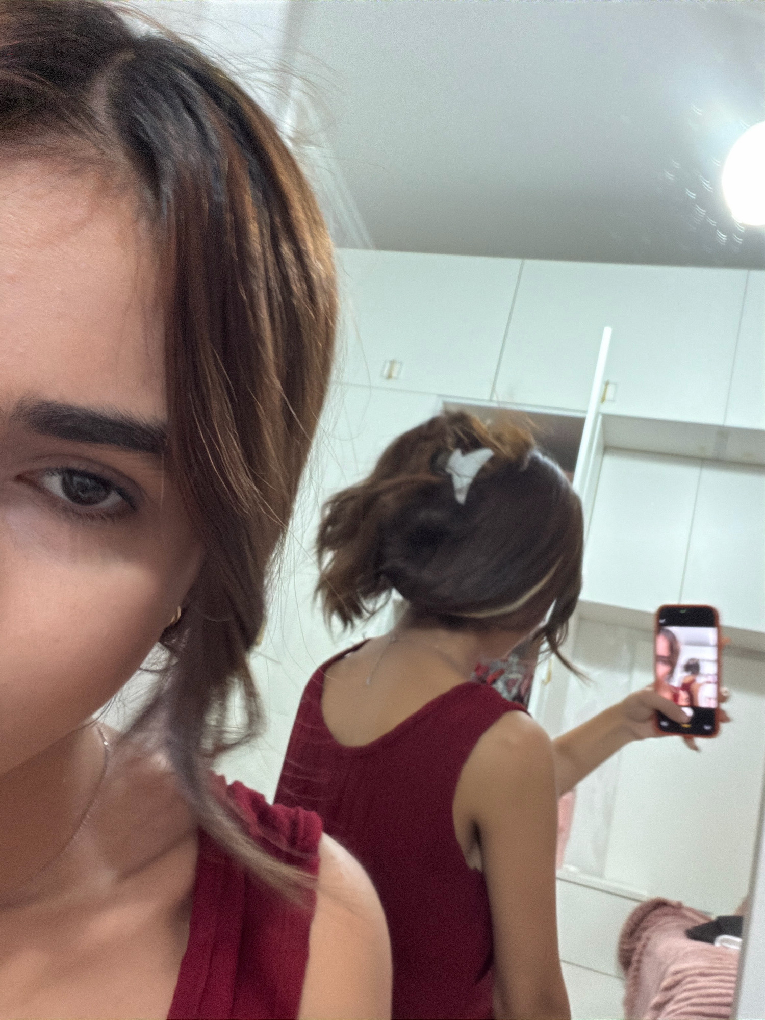 A young woman with brown hair is taking a selfie in a mirror. She is wearing a red top. The background appears to be a bedroom or dressing room with white cabinets. The woman's face is partially visible, and her left eye is looking directly at the camera. Her right arm is extended, holding a smartphone to capture her reflection. The mirror reflects the woman's back and the room behind her. There is a light fixture visible on the ceiling, casting a bright light.