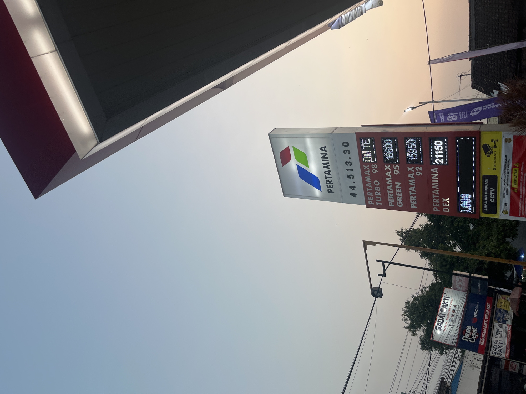 An eye-level, medium shot captures a Pertamina gas station in Boja, Indonesia, during a hazy sunset. The sky is a pale orange and blue gradient. The dominant feature is a tall Pertamina price board, illuminated by its internal lights, displaying fuel prices and types. Below the Pertamina sign, a series of other advertisements are visible, including "GADAI SAKTI," "Dana Cepat," and "Nusantara Sakti Group." A lamppost with a security camera attached stands to the left of the price board. The roof of the gas station canopy, with its red and white trim, juts into the frame from the upper right corner, casting a shadow. The overall scene is characterized by a diffused, warm light, suggesting it is either sunrise or sunset.