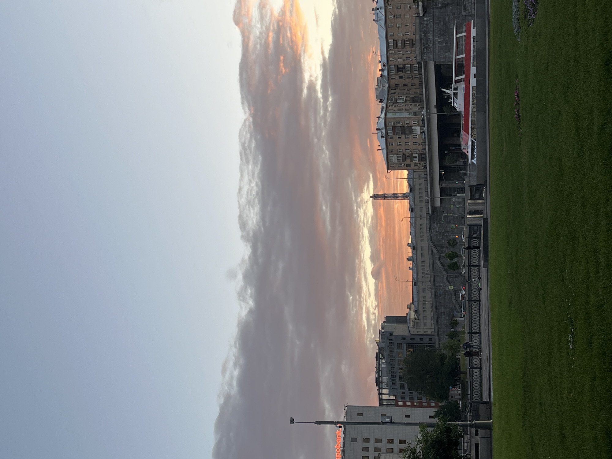 The image depicts a scenic view of the Moscow River embankment at dusk, showcasing a blend of urban architecture and natural beauty. In the foreground, a lush green lawn with scattered flowers sets a serene mood. Beyond the lawn, a pedestrian walkway is bordered by an ornate black metal fence, where two individuals are seen walking. Further along the embankment, a modern red and white passenger boat is docked, its sleek design contrasting with the older buildings that line the opposite bank.
The cityscape features a mix of architectural styles, from the monolithic structure of the Expobank building on the left, with its illuminated sign, to the residential apartment buildings with numerous windows and balconies that dominate the middle ground. A prominent industrial chimney, emitting a visible plume, stands tall against the horizon. Overarching the scene is a dramatic sky painted with hues of pink, orange, and purple, indicating a sunset, with scattered clouds adding texture and depth. The presence of streetlights and traffic lights suggests the ongoing activity of the city even as day transitions into night. The overall atmosphere is one of urban tranquility bathed in the warm glow of the setting sun.