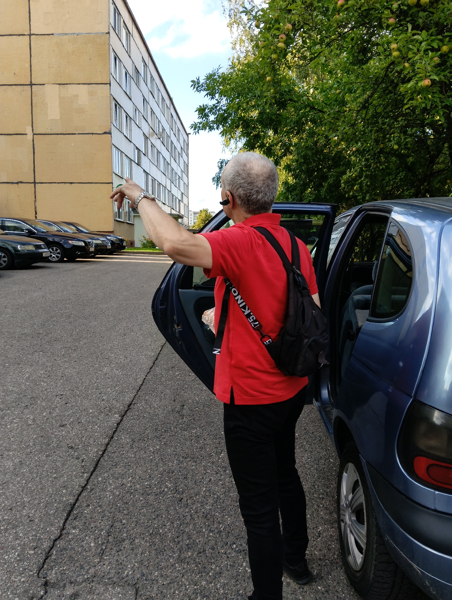 In Hrodna, Belarus, a man with graying hair, wearing a red polo shirt, black pants, and a black backpack, stands next to the open driver's side door of a blue car. He is pointing his left arm and index finger towards a multi-story residential building with a yellow facade. Several cars are parked in a row in front of the building. Trees with green leaves and visible apples are in the background. The scene appears to be a residential area or parking lot.