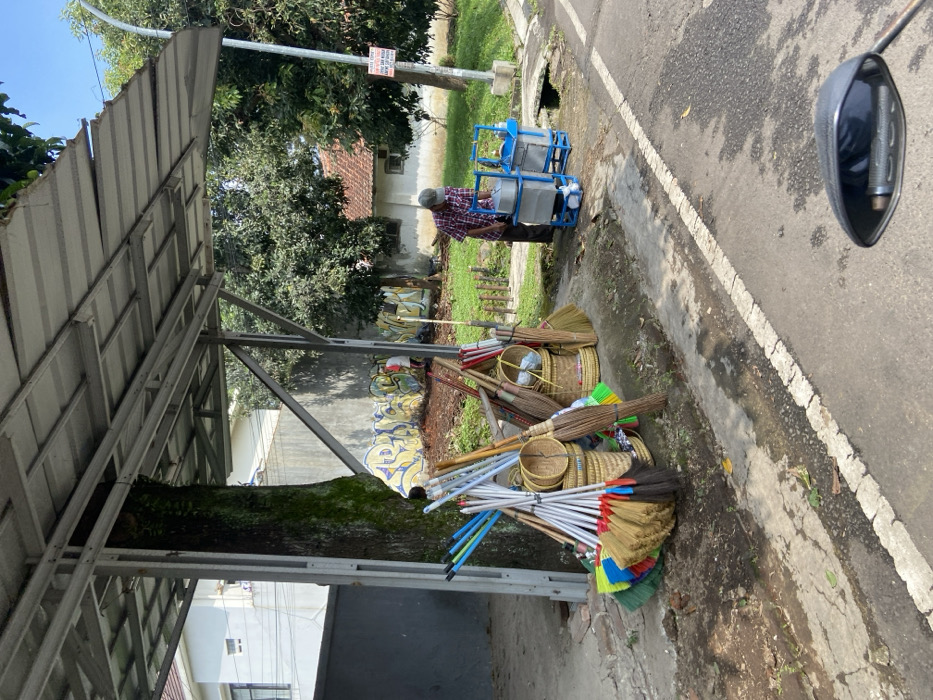 The scene is taken from the side of a paved road, looking toward a small stand selling brooms. There are 20-30 brooms for sale, arranged in two rows and resting on a concrete sidewalk. The stand is shaded by a metal awning supported by a large tree trunk. A man, wearing a blue and white patterned shirt, black shorts, and a white cap, is standing next to two blue metal containers on wheels. There is a grassy area behind the stand, and several small bushes and trees are visible. There are a few houses in the distance. The image was taken in Parongpong, Indonesia, at latitude -6.862 and longitude 107.567.