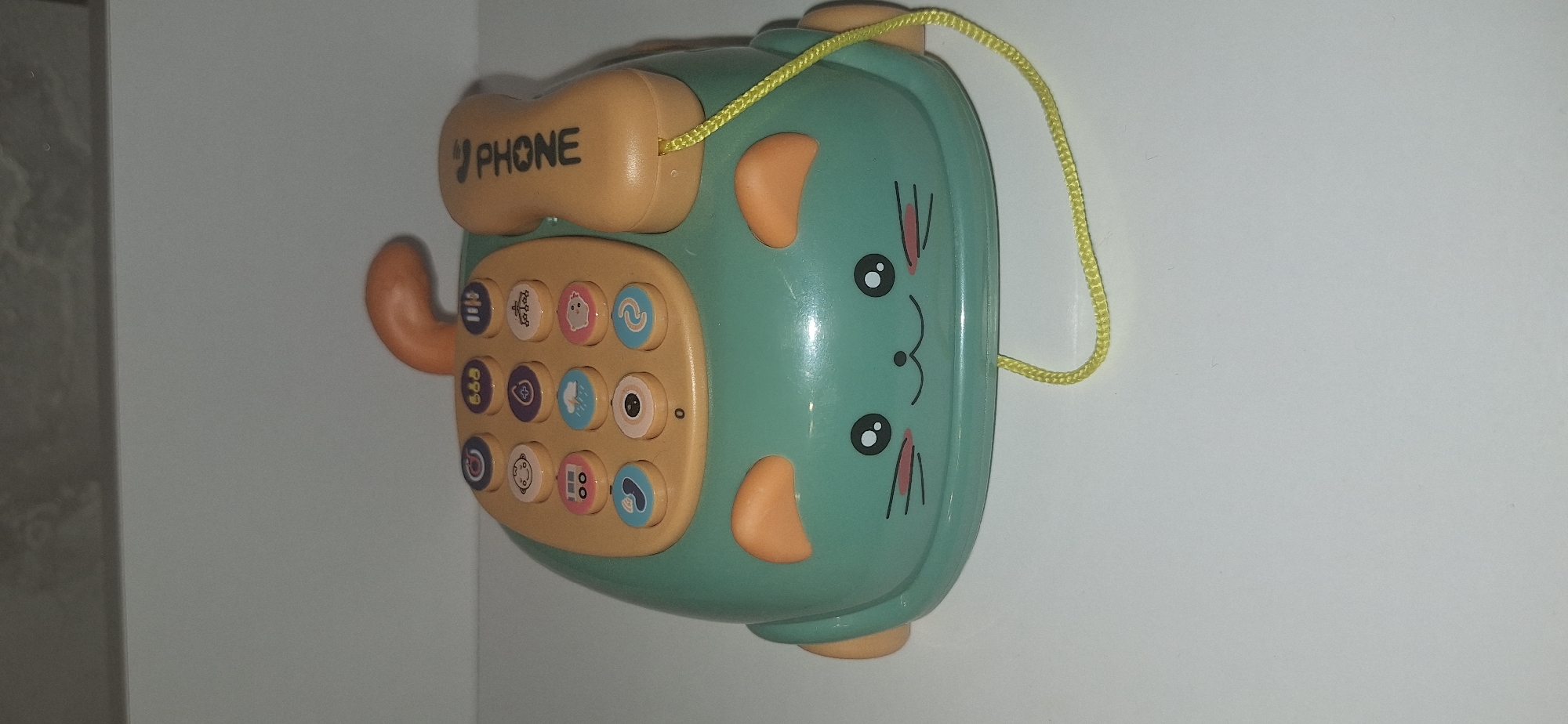 A toy telephone, shaped like a cat, sits on a white surface. The telephone is teal green with orange ears. It has a dial pad with twelve colorful buttons, each featuring a different icon. A tan-colored receiver with the word "PHONE" printed in black is attached to the side by a yellow cord. The telephone has a cute cat face drawn on the front with large eyes, a small nose, and whiskers. The background is a plain gray wall. There is no indication of the location in Bila Tserkva, Ukraine.