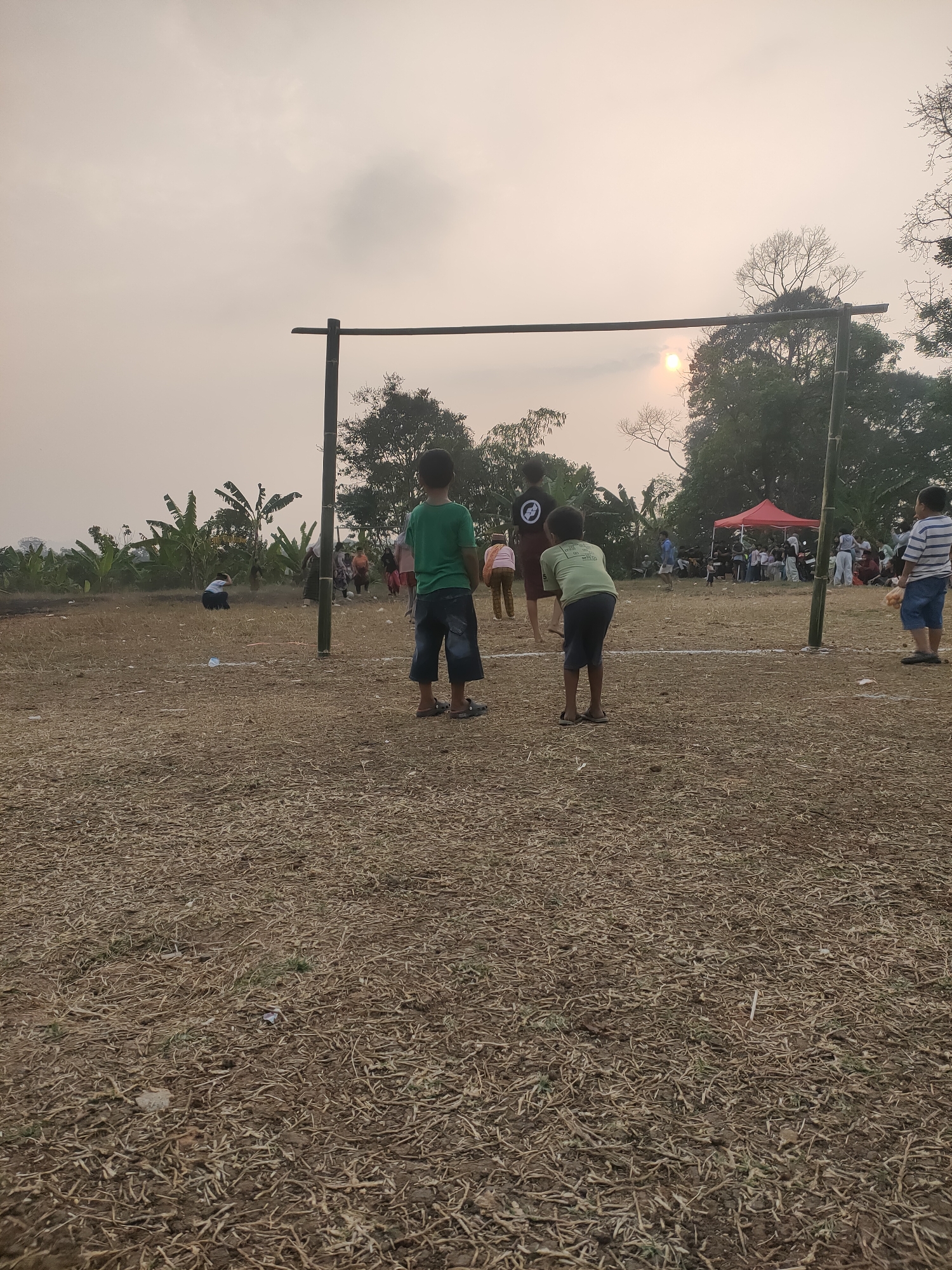 The image depicts an outdoor scene, likely in the rural outskirts of Subang, Indonesia, during what appears to be late afternoon or early evening, given the hazy sunlight filtering through the overcast sky. The foreground is dominated by dry, brown, straw-like ground cover, suggesting a field that has recently been harvested or is arid.
In the mid-ground, a makeshift goalpost constructed from bamboo poles stands prominently, with one vertical pole slightly off-center to the right. Two young boys are visible near this structure. The boy on the left, facing away from the camera, wears a green t-shirt and dark shorts. To his right, another boy, also facing away, wears a light-colored t-shirt and dark shorts. A few other figures are scattered throughout this area, some indistinct.
Further in the background, a red tent is set up, and a group of people are gathered around it. Banana trees are visible on the left side of the image, indicating a tropical or subtropical environment. The background also shows a silhouette of trees against the sky and a distant, hilly landscape.
In the far right of the image, a young boy wearing a striped t-shirt and blue shorts stands, looking towards the group near the tent. The overall impression is of a casual community gathering or event taking place in a rural setting.