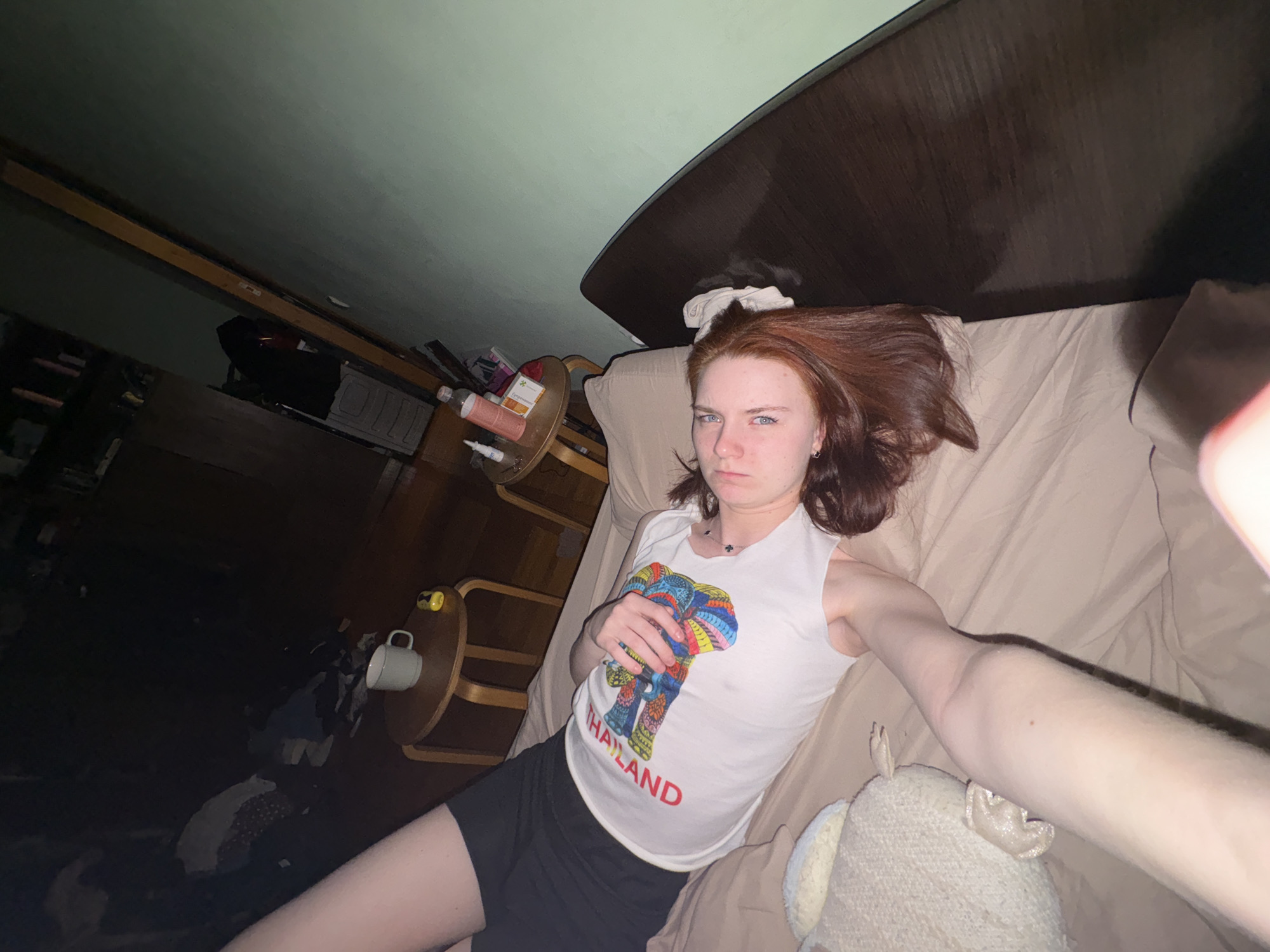 A young adult with reddish-brown hair lies on a tan bed in a dimly lit room. She is wearing a white tank top with a colorful elephant print that says "THAILAND" in red lettering, and black shorts. To her left, a nightstand holds a mug and other items, and beyond that, the floor is cluttered. A teddy bear rests near her on the bed. The background is dark and indistinct, obscuring details of the room. The location in the city of Unknown, Unknown is unknown.