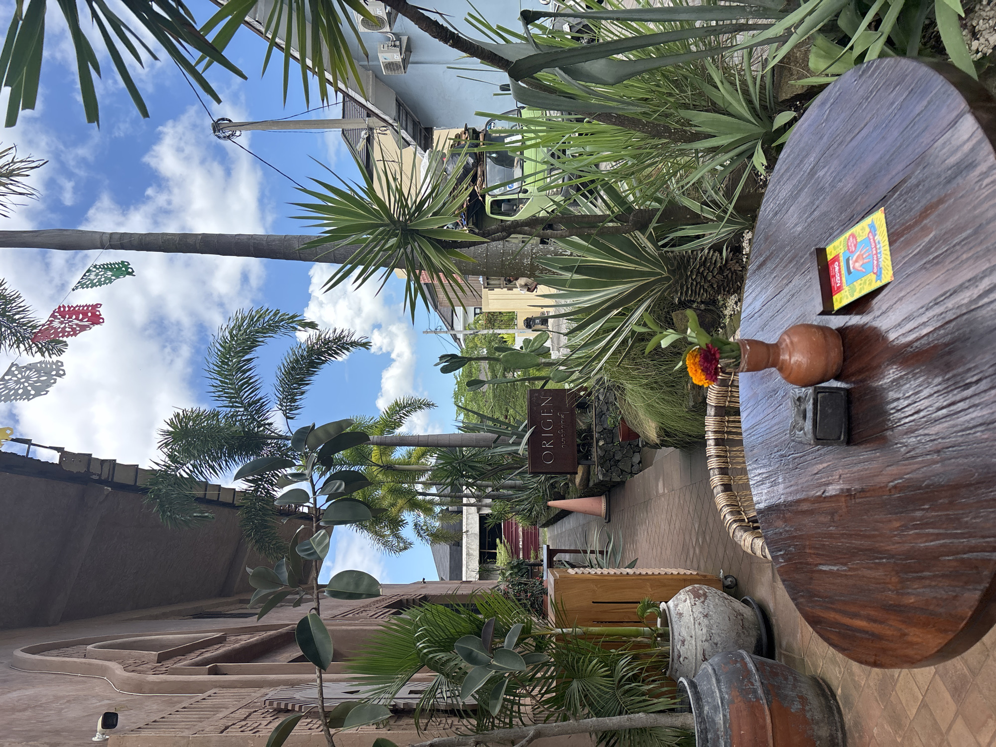 The image captures a partial view of the exterior of a business named Origen, located in Mengwi, Indonesia. In the foreground, a round wooden table sits prominently, adorned with a small vase containing a flower and a box. A small black object is also present on the table. The table appears to be placed beside a walkway adorned with pots. Various types of greenery, including palm trees and other plants, are dispersed throughout the scene. In the background, there is an overcast blue sky. A street with other buildings is visible in the distance, with utility poles and wires. Decorations and structures are located along the building.