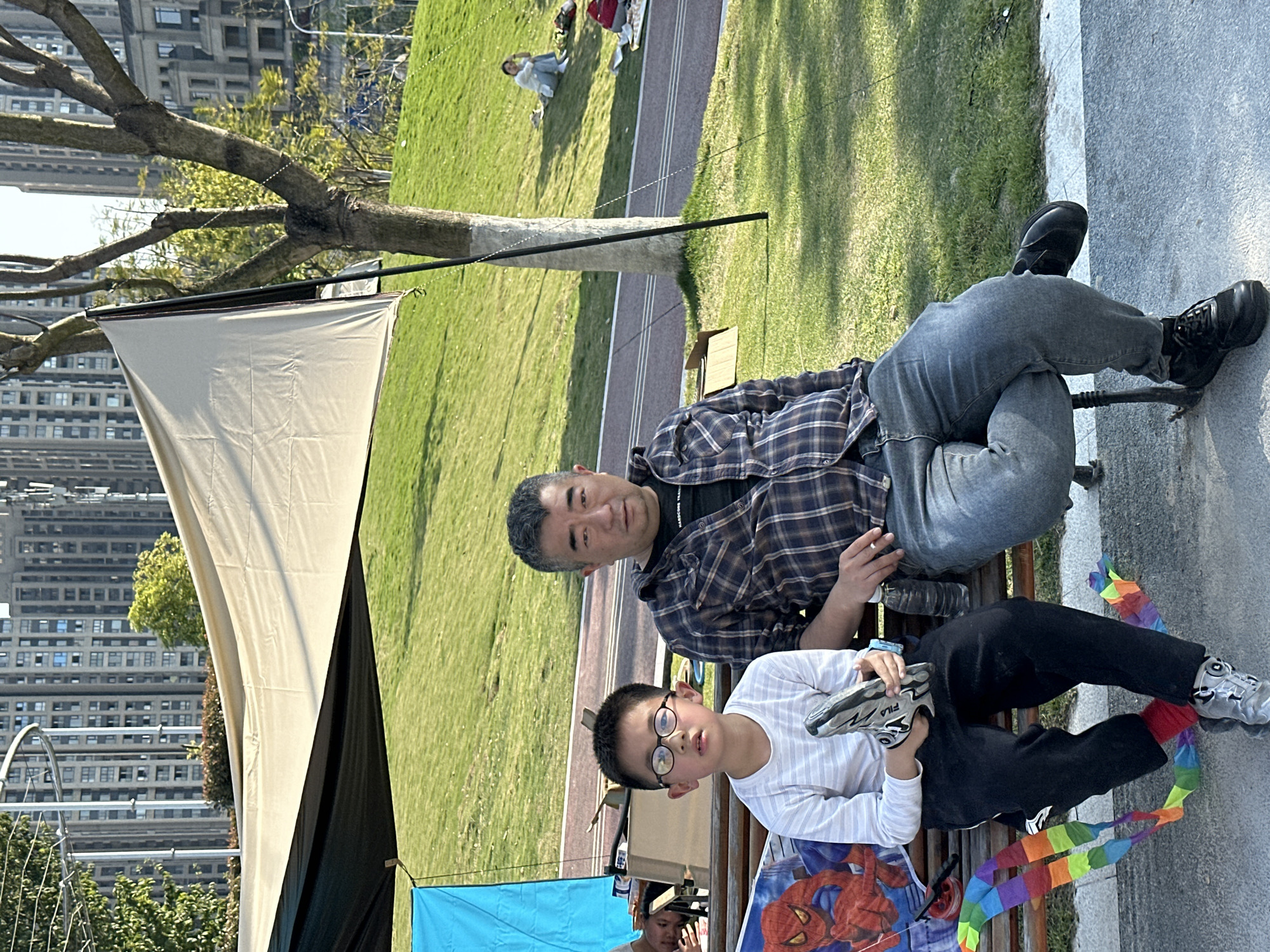 A man and a boy are sitting on a bench in a park on a sunny day. The man is wearing a plaid shirt and jeans, and the boy is wearing glasses, a striped shirt, and black pants. The boy is holding a pair of shoes, and there is a colorful kite on the bench next to him. In the background, there is a grassy field, a walkway, a tree, a tent, and a cityscape of tall buildings. The location is Jiangjin, China.