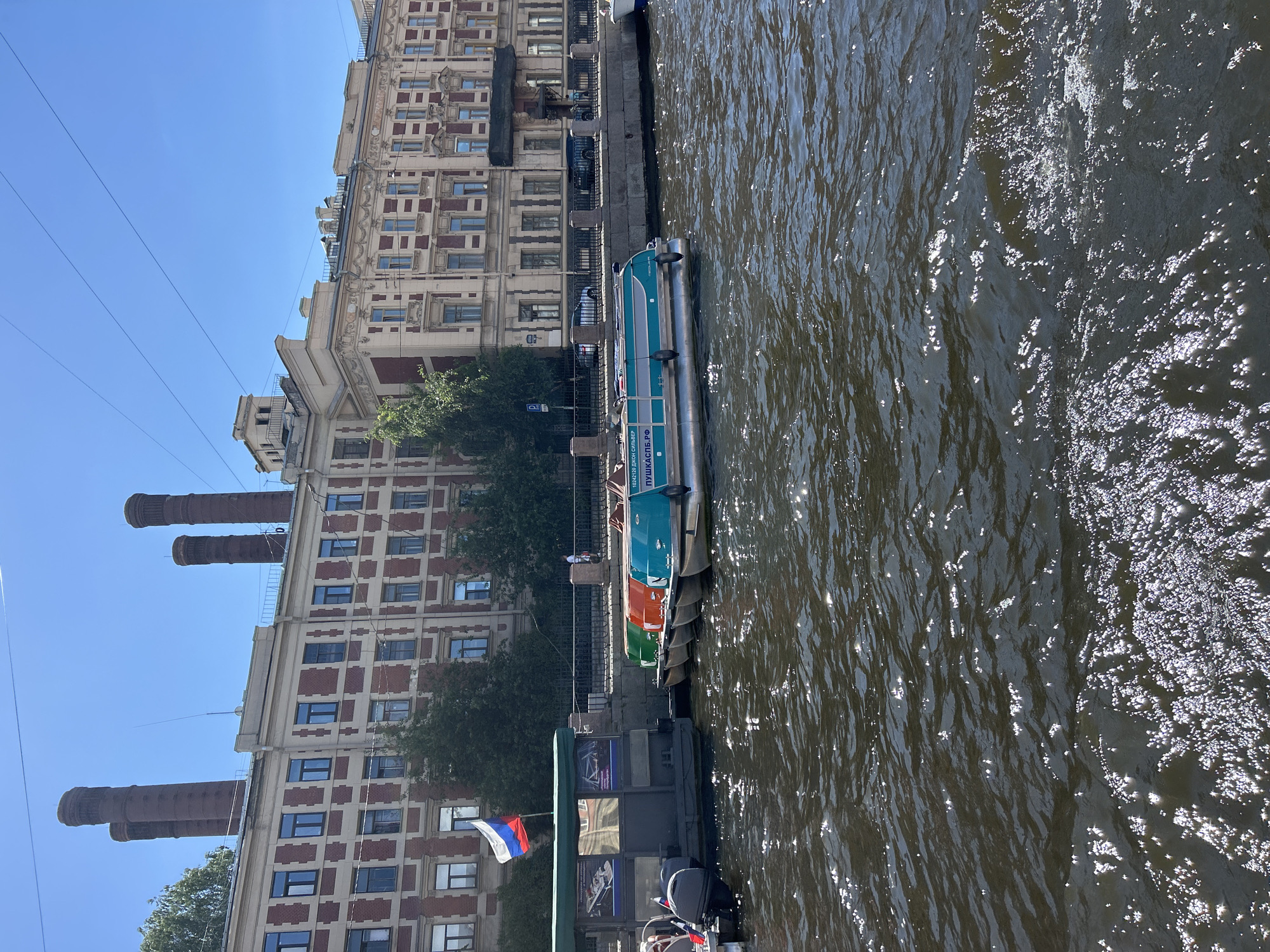 The image depicts a canal in an urban environment with boats docked along the bank. A large brick building with multiple windows and four prominent chimneys stands in the background. A Russian flag is visible on a structure next to the canal. Several small boats, including a turquoise boat with Cyrillic lettering, are moored. The water is dark and choppy, reflecting sunlight. The sky is clear and blue.