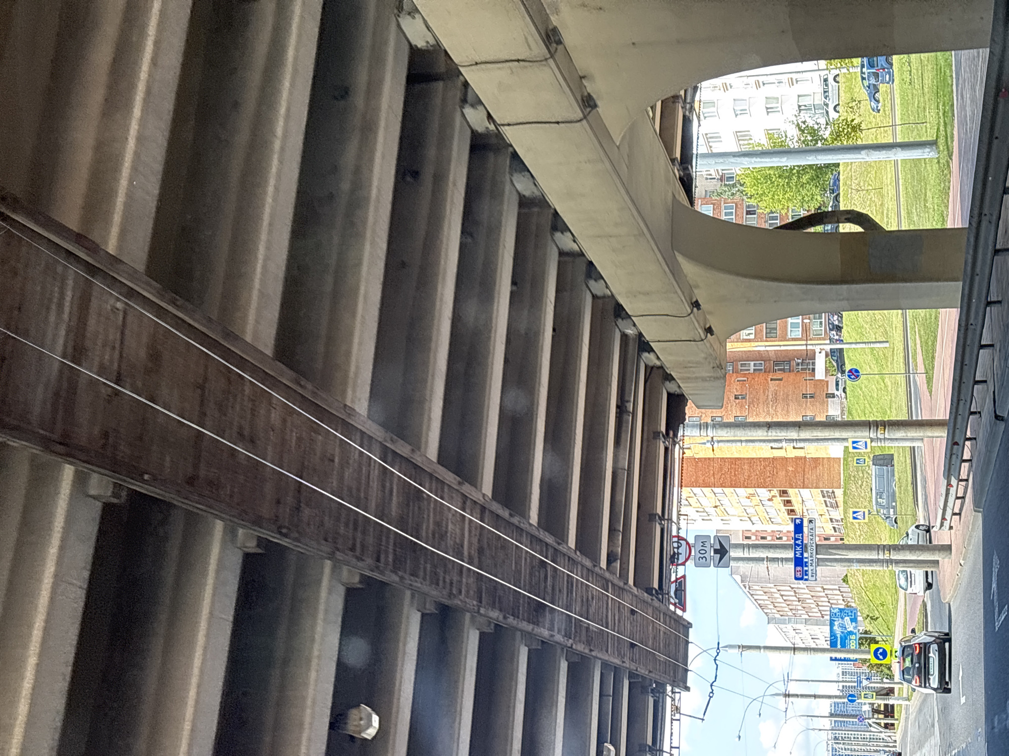 The image shows an underpass in Minsk, Belarus, likely on the MKAD ring road, given the sign indicating direction to the MKAD. The perspective is from a vehicle, looking upwards at the concrete structure of the overpass. Two parallel wooden beams run horizontally along the underside of the overpass, suspended by wires. These beams appear to be aged and weathered, with visible wood grain and some signs of wear. Below the overpass, a street scene is visible, with cars, streetlights, and buildings in the background. One car, a white BMW i3, is in the foreground, and other vehicles are parked or driving in the distance. Several traffic signs are also visible, including speed limit signs and directional signs. The overall scene suggests an urban environment with infrastructure for transportation.