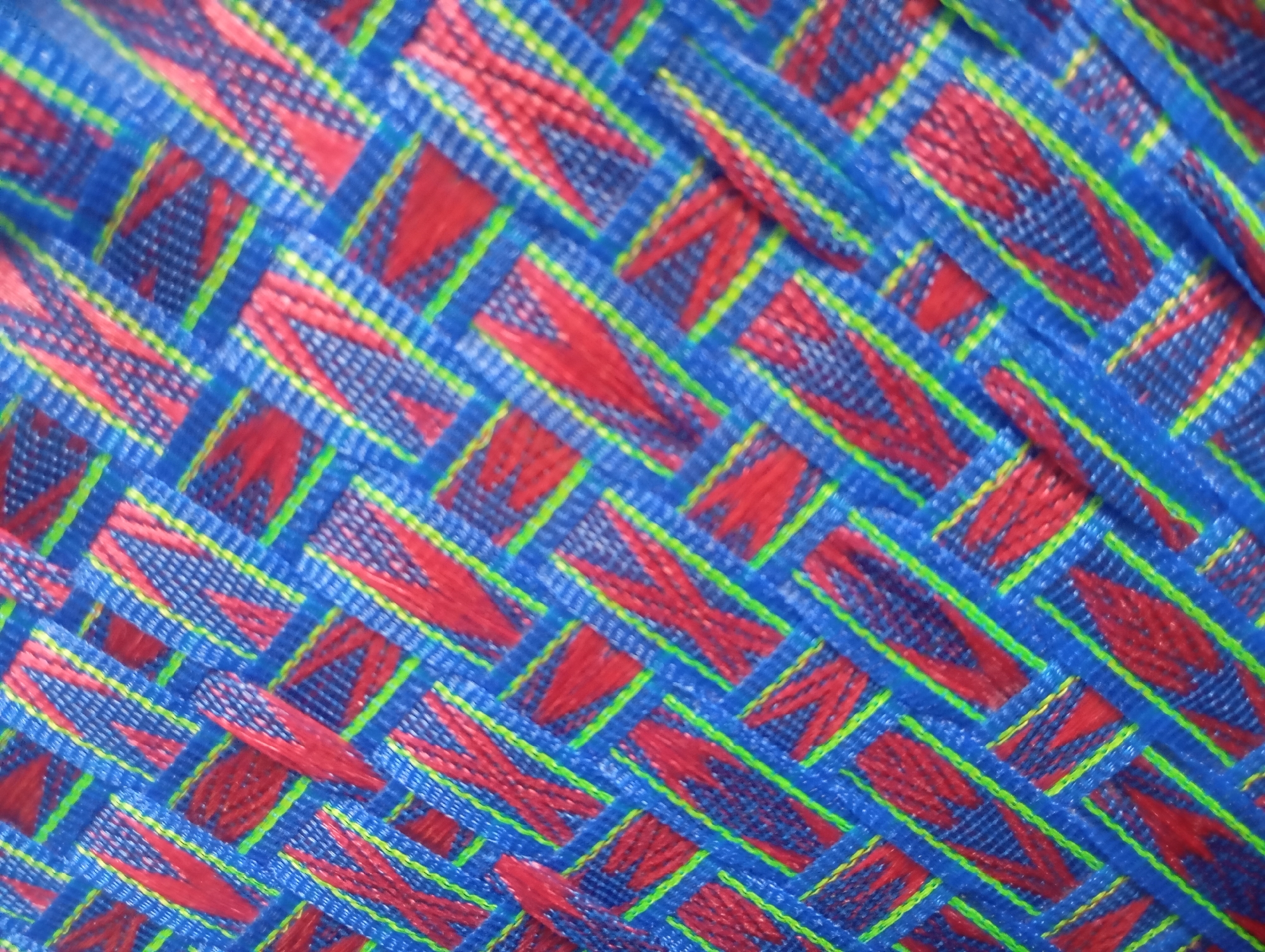 A tightly woven fabric features a repeating pattern of diagonal red elements interlaced with blue and thin green threads. The fabric appears to be synthetic and is viewed from a top-down perspective, creating an abstract visual texture. The lighting is even, with no discernible shadows or highlights. The location is unknown.