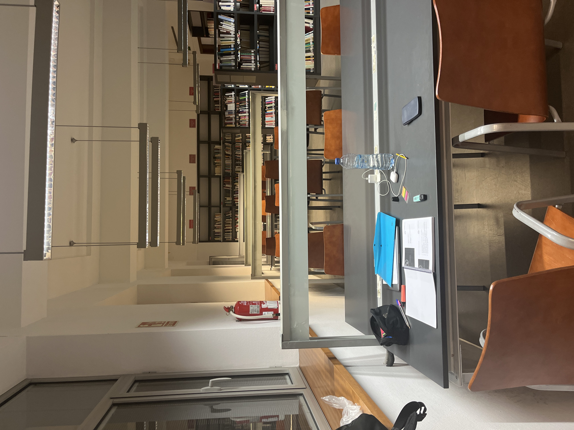 The image shows the interior of a library. Long rows of bookshelves, filled with books, extend into the distance. Rows of tables and chairs are arranged between the bookshelves. On one table, there's a water bottle, phone, papers and a blue folder. A fire extinguisher is visible on the wall. The location is likely a library in València, Spain.