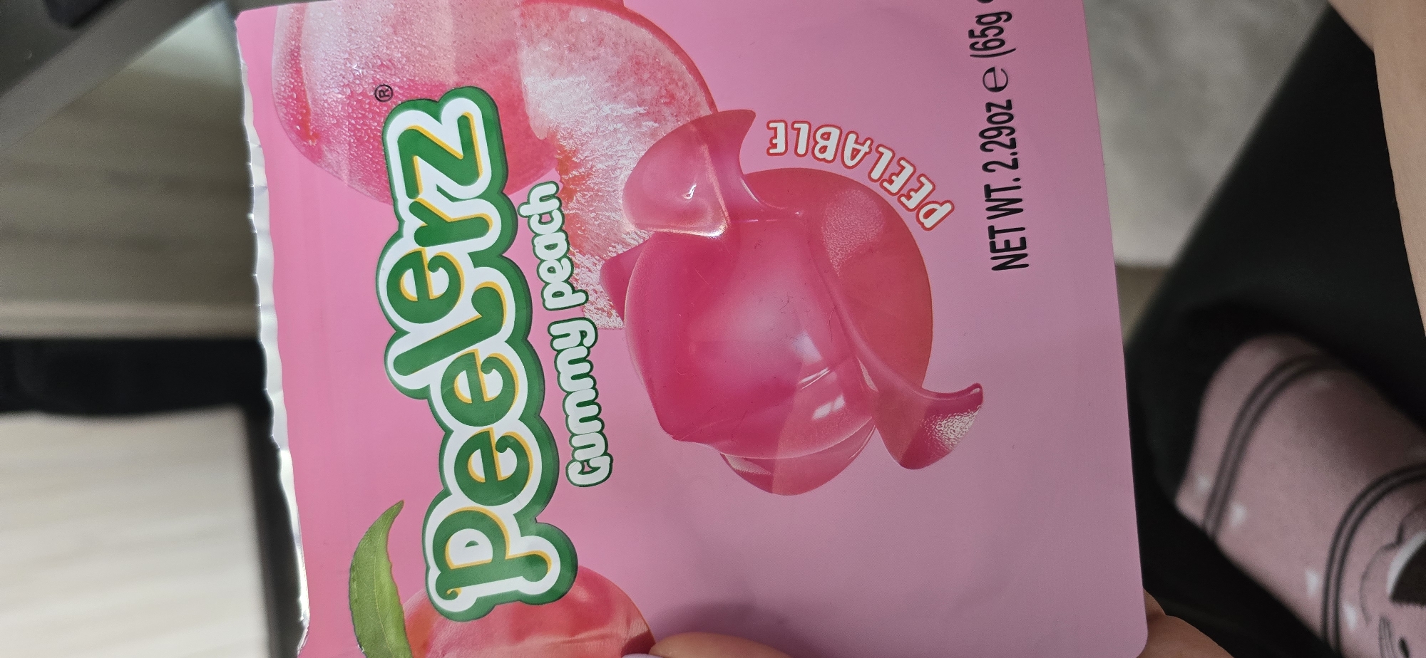 The image shows a package of "Peelerz Gummy Peach" candy. The packaging is pink and features images of a peach and a gummy peach candy. The words "Peelerz Gummy Peach" are printed in green lettering. The text "PEELABLE" and "NET WT. 2.29oz e (65g)" is also visible. Parts of a black chair, the edge of a white surface, and a person's clothing are also visible in the frame. The image does not provide any information about the location in Burgas, Bulgaria.