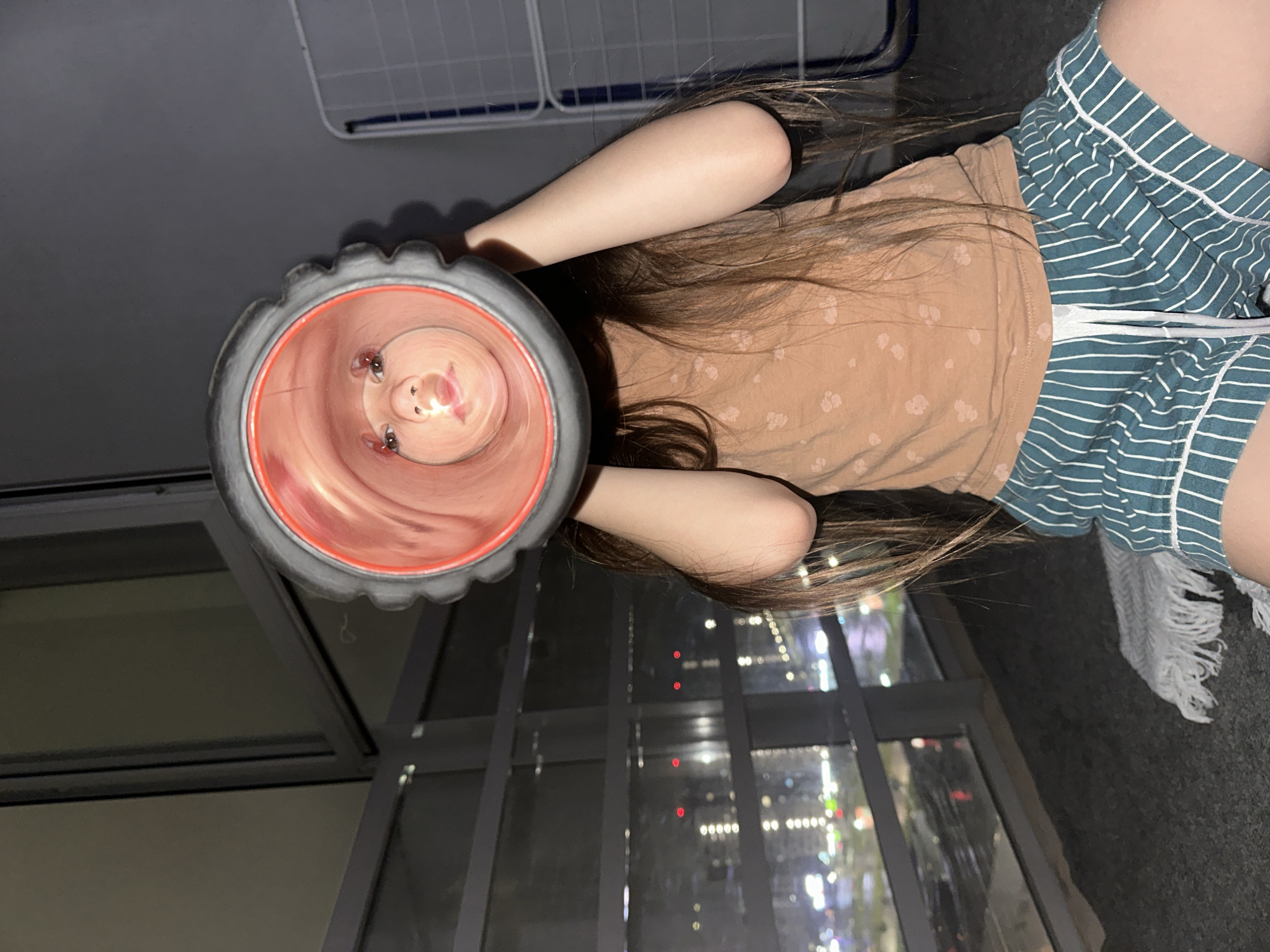 The image depicts a young child indoors at night, described as taking place in Izhevsk, Russia. The child, with long, dark brown hair, is kneeling or sitting on a dark gray carpet. They are wearing a light brown short-sleeved top with a subtle, lighter-toned print, and greenish-blue shorts featuring white vertical stripes and a white drawstring.
The child holds a dark gray, textured cylindrical object, possibly a foam roller or a large toy, with a prominent red interior rim, directly to their face. The child's eyes, nose, and mouth are visible and distorted by the perspective inside the cylinder, illuminated by light reflecting into the object. Their arms are bent, holding the object aloft.
To the immediate left of the child's knee, a white, loosely woven or knitted throw blanket is partially visible on the carpet. On the far right, a segment of a white, rectangular gridded rack, possibly a clothes drying rack, is visible against a plain light gray wall.
Behind the child, a large window offers a panoramic night view of a city. Numerous illuminated buildings, streetlights, and vehicle lights (including distinct red and white points of light) are visible, reflecting off wet surfaces, suggesting recent rain or a damp environment. Dark window frames segment the exterior view.