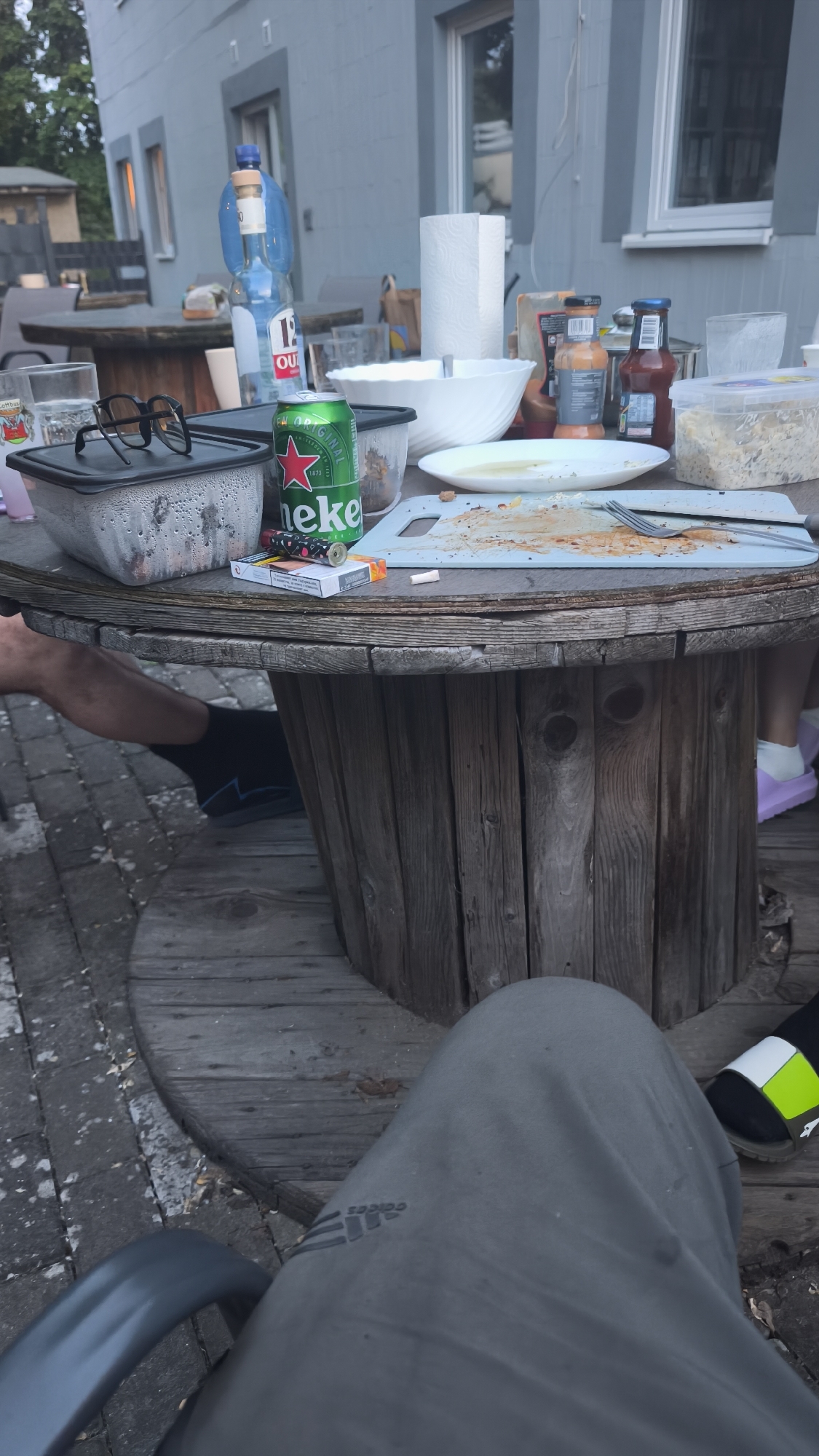 This image captures an outdoor casual gathering during dusk in the city of Unknown, Unknown. The central object is a large, weathered wooden cable spool repurposed as a table.
On the table, various items indicate a recently concluded meal. In the foreground, a rectangular plastic container with a dark lid, showing condensation, sits next to a green Heineken beer can. A pair of black sunglasses rests on the container's lid. To the right of the beer can, a pack of cigarettes and a lighter are visible, with a single loose cigarette lying on the table. Further right, a light blue cutting board is covered with food residue, and a fork and knife are placed upon it. Behind the cutting board, a white plate with residue, a white ceramic bowl, and a roll of paper towels are arranged. Several bottles of condiments, including ketchup and an orange sauce, are also present. A clear plastic container containing food, possibly salad, is positioned on the far right. In the background, a large plastic water bottle and a smaller glass bottle with a cork are visible.
Three individuals are partially visible. In the bottom left, a person's bare leg and a foot wearing a dark sock are seen under the table. In the bottom right, another person's leg and foot, clad in a light purple sandal, are visible. Dominating the bottom center is the lap of a third person, wearing grey sweatpants with an Adidas logo, seated in a chair whose arm is partially visible on the left.
The ground surrounding the table consists of grey interlocking pavers on the left, transitioning to wooden decking under the table. In the background, a two-story grey building with multiple windows is visible. The lighting suggests late afternoon or early evening.