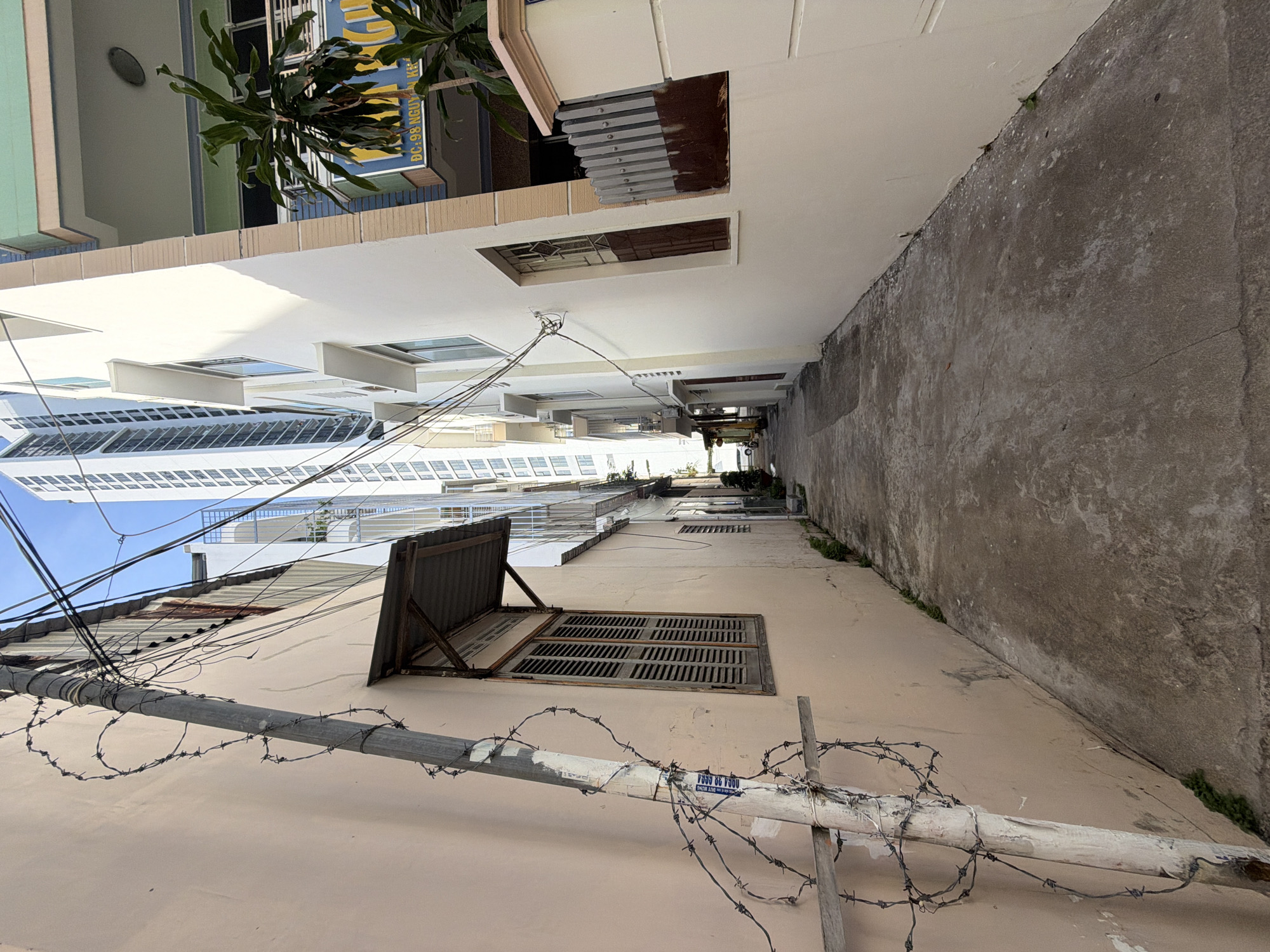This image captures a narrow alleyway in the Bac Nha Trang Ward of Vietnam, flanked by buildings on either side. The alley is paved with concrete and shows signs of wear and tear, with cracks and discoloration. Barbed wire is coiled around a utility pole on the left, suggesting a security measure. The building on the left has a pale pink facade and a wooden-shuttered window. The building on the right is white, with multiple windows and balconies. In the background, a tall, modern skyscraper stands out against the clear blue sky, contrasting with the older, narrower buildings. The alley appears to be a service entrance or a less-trafficked area. There are no people visible in the immediate scene.