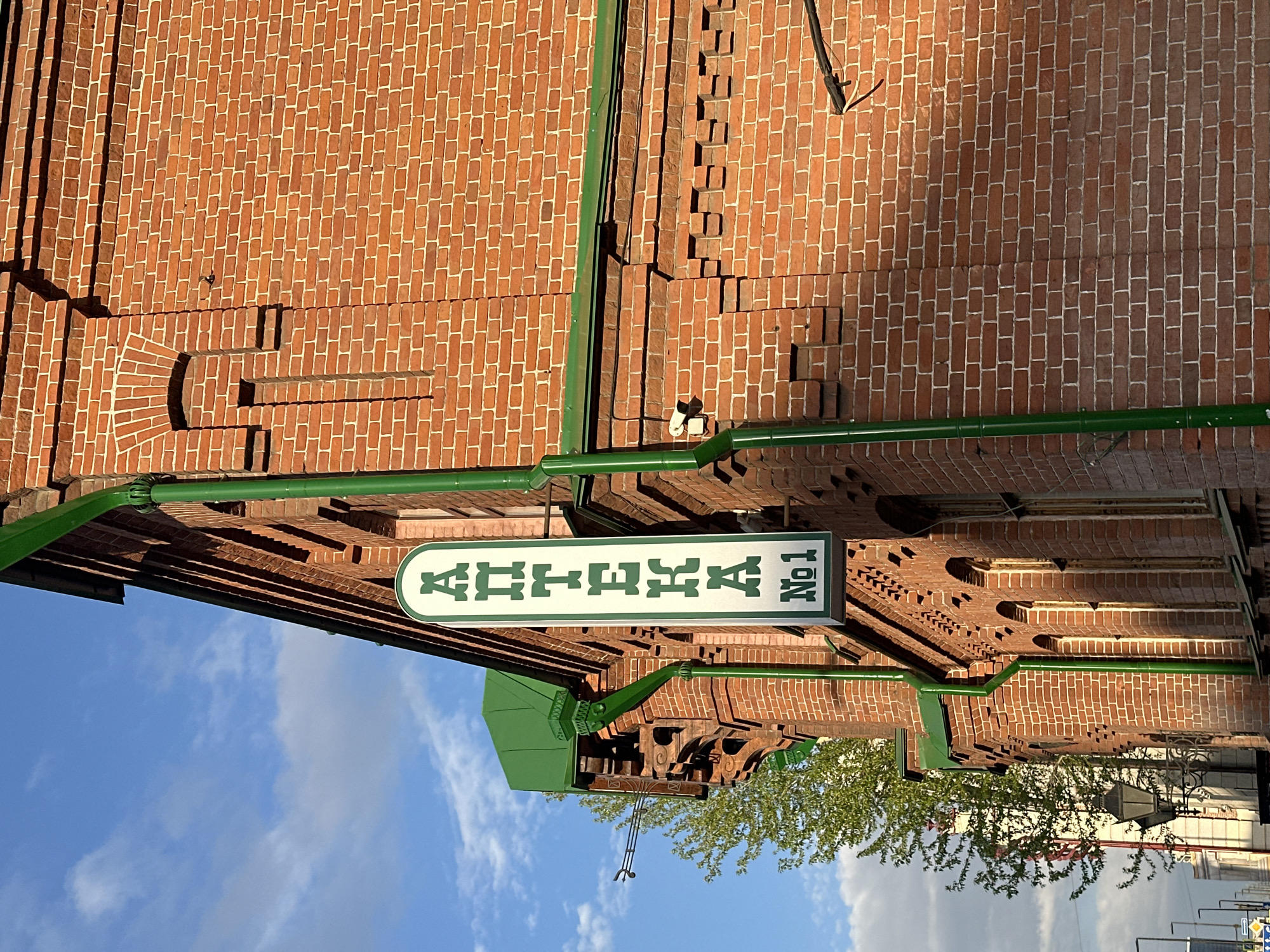 The image shows the "Aptheka No.1" sign, a historical pharmacy in Krasnoyarsk, Russia. The building is constructed of red brick. The sign is rectangular with rounded ends, white in color, and displays the name "Aptheka No.1" in green, written in the Cyrillic alphabet. Green pipes are attached to the building near the sign. The background includes a blue sky with clouds and the top of a tree.