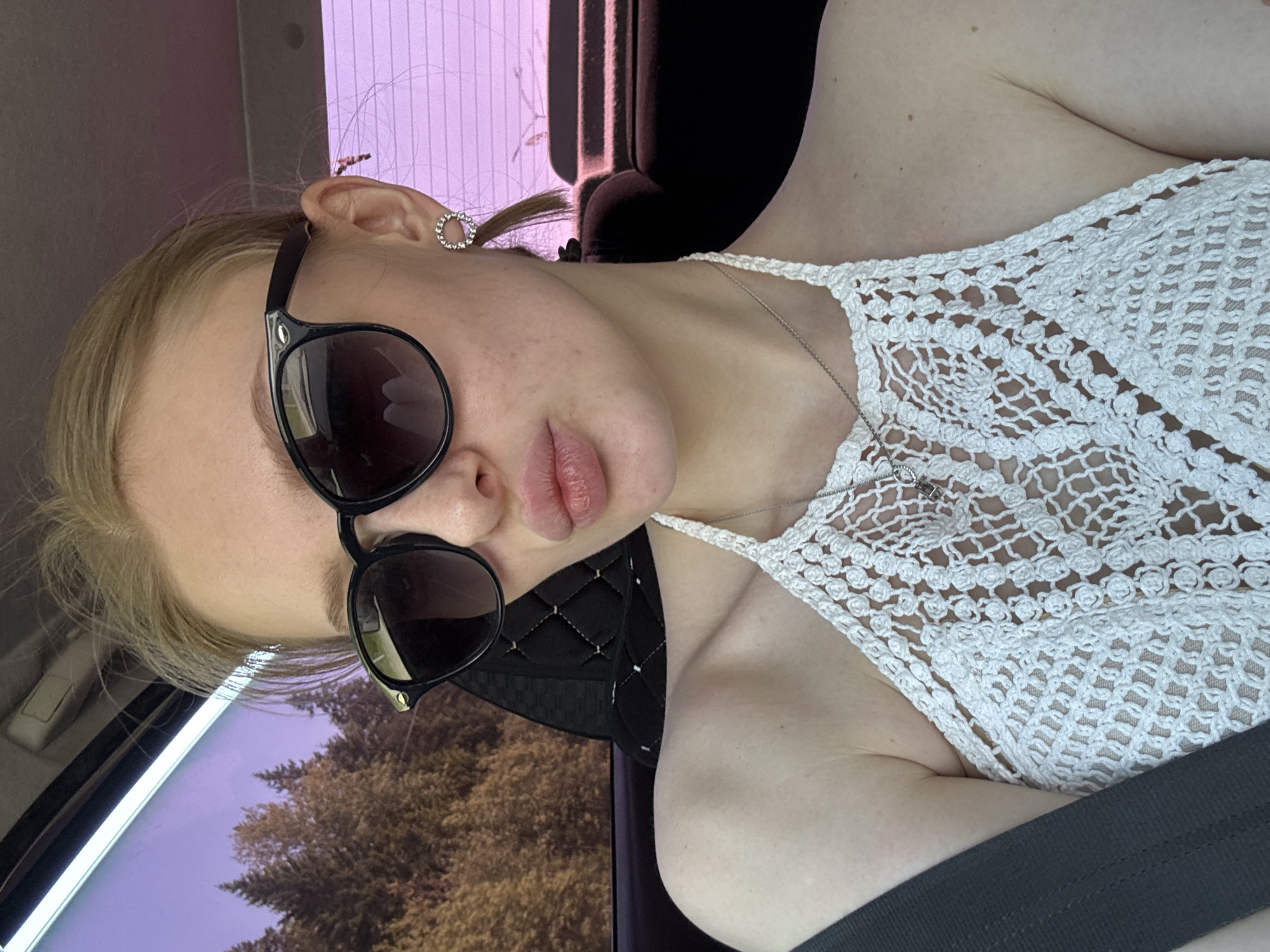 A young woman is positioned in a car. She is wearing sunglasses and a white crocheted top with a silver necklace. The car's window is visible, with a view of trees outside, possibly in a wooded area. She is wearing a gray seatbelt. She is wearing a circular earring in her left ear. There is no explicit indication of Perm, Russia in the image.