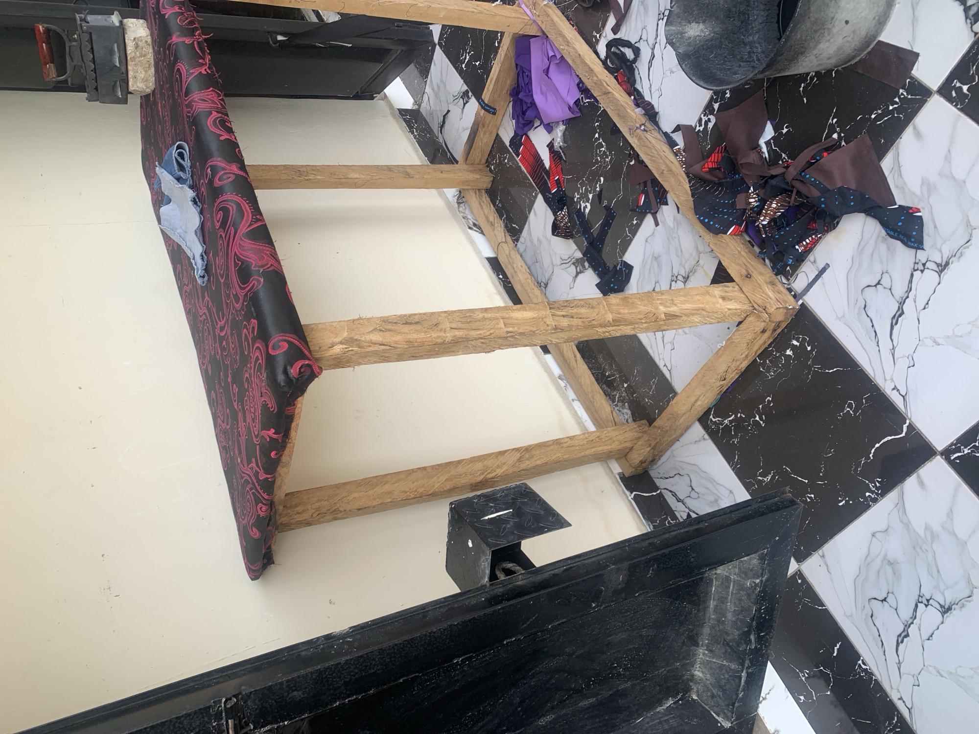 In Jauro Yinu, Nigeria, a makeshift ironing board stands outside a building. The board has a black fabric with a pink paisley pattern and is supported by rough wooden legs. Various colorful fabric scraps are scattered around the base of the ironing board and on the checkered floor. A black metal object is attached to the wall to the left of the ironing board.