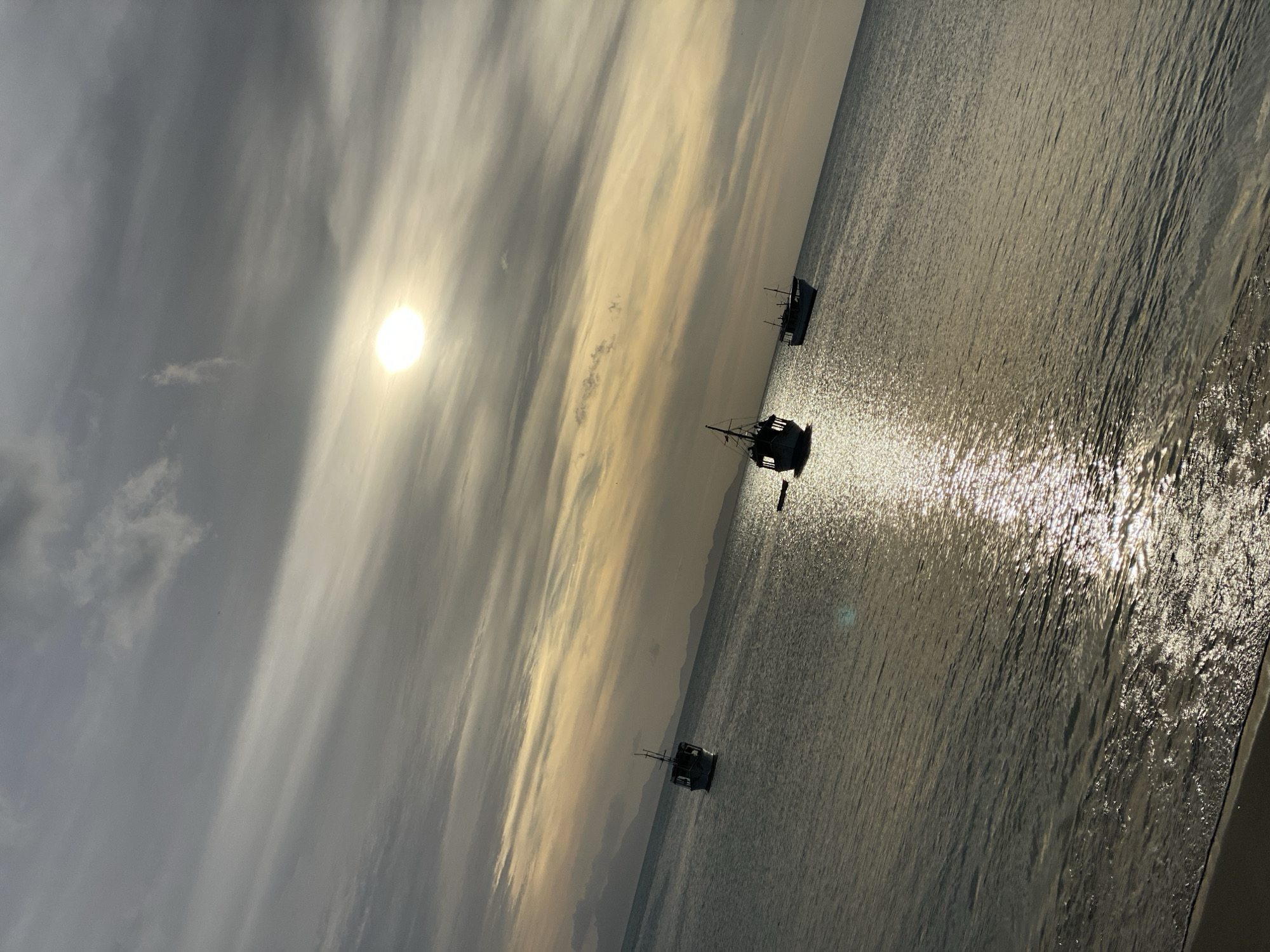 The image displays a coastal scene in Juan Griego, Venezuela, captured during a sunrise or sunset. The upper portion features a sky filled with stratus and altostratus clouds, presenting a gradient of gray, yellow, and orange hues. A prominent, bright circular sun is positioned in the upper center-right, partially veiled by these clouds. Below the sky, a calm body of water, indicative of the sea, occupies the majority of the frame. This water surface exhibits a strong specular reflection of the sun, creating a shimmering path that extends from the horizon towards the bottom center of the image. Three dark-silhouetted fishing boats are observed stationary on the water. One boat is positioned on the far left, further offshore. A second boat is located centrally, closer to the viewer, displaying a discernible cabin and mast. A third boat is partially visible along the far right edge of the frame. A narrow strip of sandy shoreline with minor breaking waves is present in the bottom-left corner. Faint, dark outlines of distant landmasses are discernible along the horizon line. No people are visible within the scene.