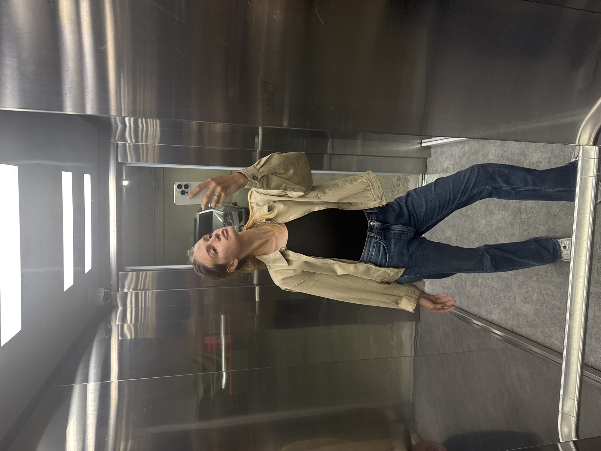 A person stands in an elevator, taking a picture in the mirror. The individual is wearing blue jeans, a black top, and a tan jacket. The elevator has stainless steel walls and a gray floor. The location is Unknown, Unknown.