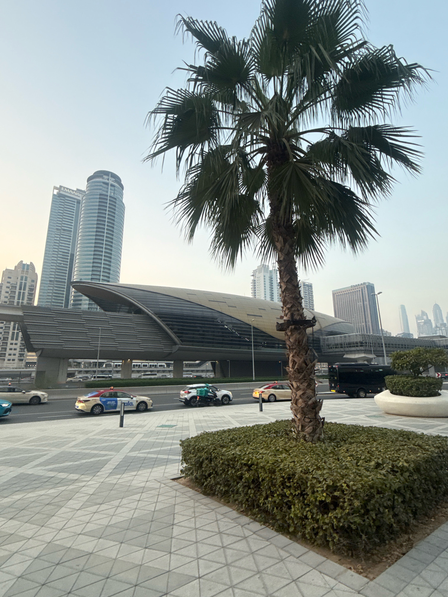 The image depicts a street scene in Dubai, United Arab Emirates, likely near the Dubai Marina or Jumeirah Beach Residence area, given the modern architecture and palm trees. In the foreground, a tall palm tree stands in a square planter filled with low green bushes. The ground is paved with geometric patterned tiles. To the left, a modern metro station with a distinctive curved roof is visible. Cars are driving on a road in front of the metro station. Among the cars, a white taxi with a red stripe is prominent. In the background, several tall, modern skyscrapers dominate the skyline, including the distinctive Emaar buildings, suggesting a central business or residential district. The sky is a pale, hazy blue, indicating either early morning or late afternoon light. The scene is devoid of any visible people.