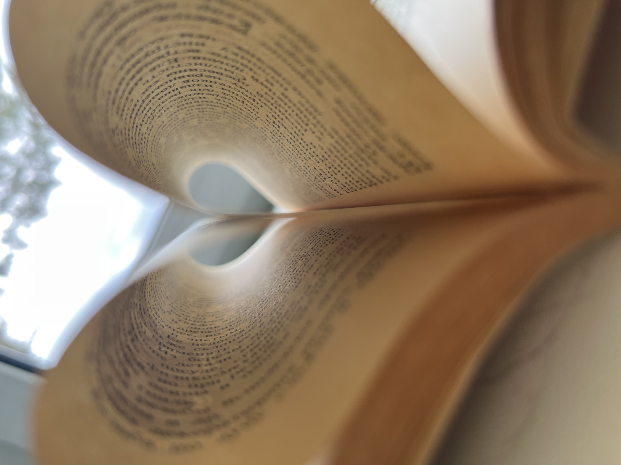 A close-up shot focuses on an open book, its pages folded back to create a heart-like shape. The pages, aged and yellowed, are filled with dense black text. A bright, blurred light enters from the left side, likely from a window. The location is suggested to be Chaikovsky, Russia, though this cannot be confirmed from the image alone. There are no people in the image.