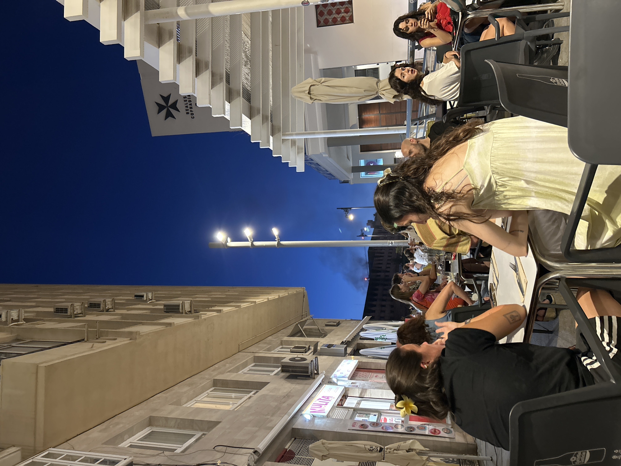 This media file captures an evening scene at an outdoor dining area in Málaga, Spain. The image is rotated, showing the deep blue twilight sky at the top. Numerous people are seated at dark tables and chairs, engaged in dining and socializing.
In the foreground, a person with long dark hair and a yellow flower tucked behind their ear wears a black top and striped shorts, facing away from the camera. Nearby, another person in a light yellow-green dress leans over a table. Throughout the mid-ground and background, other diners can be seen, some looking towards the camera, others conversing or looking at their phones.
The setting includes multi-story buildings, some with visible windows and air conditioning units. On the left, a prominent white pergola structure extends over a section of the outdoor seating. Beneath this pergola, a white sign bears a black Maltese cross symbol and the text "REALES COFRADIA." Further down the street, an illuminated sign displays the word "MULITA." Streetlights are visible, casting light on the scene, indicating it's nighttime. A folded beige parasol stands near some tables. The atmosphere appears lively and relaxed, typical of an evening out in a city. The back of one chair clearly shows the outline of a Coca-Cola bottle.
