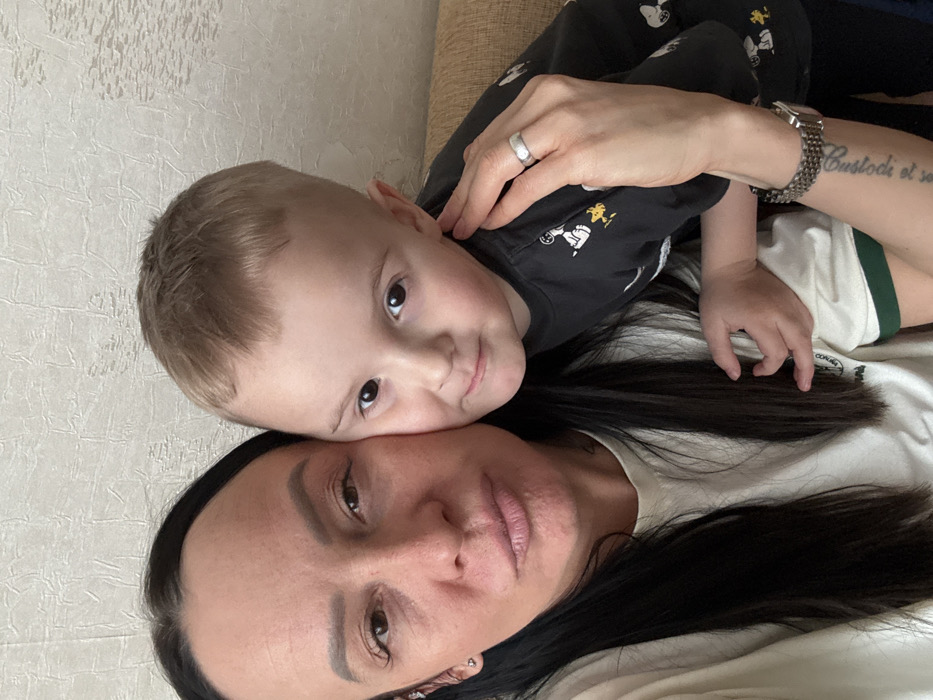 An indoor image taken in Pavlovsky, Russia, displays an adult female and a young male child. The female subject, positioned on the left, has long dark hair, a light complexion, and her mouth slightly pursed. She wears a light-colored t-shirt with a green collar stripe. A silver stud earring is visible in her left ear, and a tattoo with vertical script is on her left forearm. A silver-toned watch with a metal link band is on her left wrist, and a plain silver-toned ring is on her left ring finger. The child, a young boy with short blond hair, is positioned on the right, leaning his head against the adult female's right cheek and shoulder. He wears a dark long-sleeved shirt adorned with multiple white and yellow cartoon characters. Both individuals look directly towards the viewer; the child exhibits a subtle smile. The adult female's left hand rests on the child's left shoulder, and his right arm is around her back. The background features a light-colored wall with a textured, irregular pattern and a visible portion of a light brown upholstered surface on the far right.