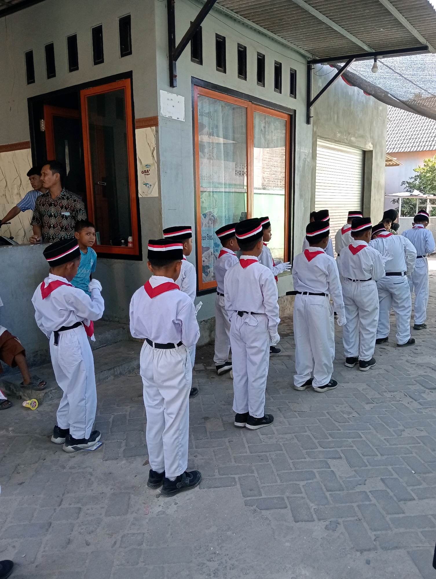 In front of a building with a gray facade and orange-framed windows, a group of children in white uniforms with red neckerchiefs and black hats stand in formation on a brick-paved area. The children appear to be participating in a parade or similar event. Two adult men stand near an open doorway, one wearing a patterned shirt and the other a striped shirt. The building's roof is covered with corrugated metal and netting. The scene appears to be outdoors in a village or suburban area. The location is Pagerungan Kecil, Indonesia.