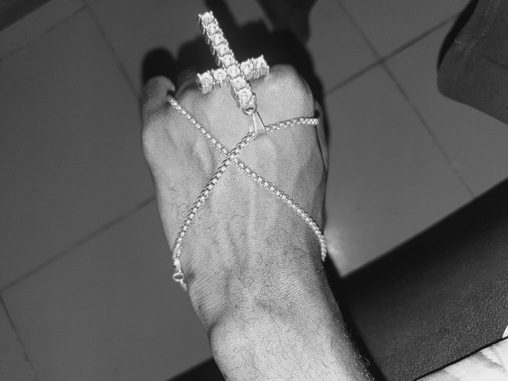 A black and white close-up image shows a person's hand adorned with jewelry. A ring in the shape of a cross, studded with what appear to be gemstones, is worn on the hand. A delicate chain wraps around the hand, crossing over the back of the hand from the wrist to the base of the fingers. The background is a tiled floor. There is no information in the image to suggest the location is Lokoja, Nigeria.