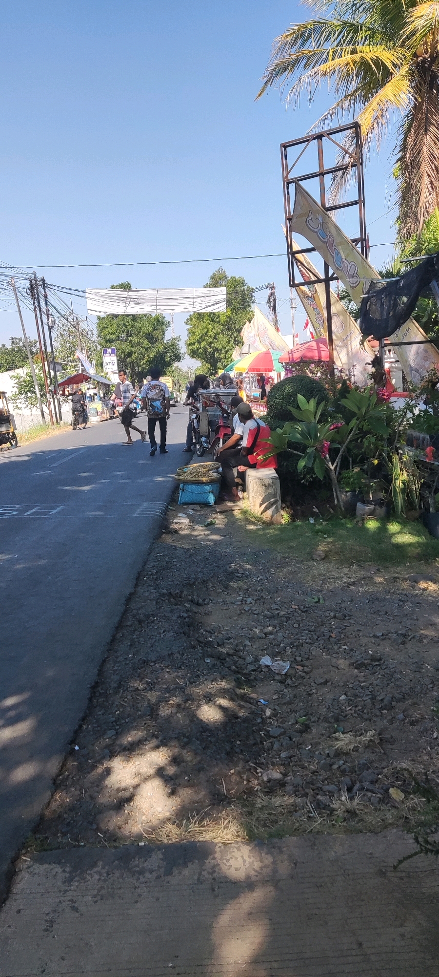 The image depicts a street scene in Belawa, Indonesia, on a bright, sunny day. The road is asphalted, with a gravelly shoulder on the right side. Several people are present, some walking on the road, others gathered near stalls. A few motorcycles are parked on the side of the road.
In the foreground, on the right shoulder, a person wearing a red shirt and black pants is seated on a concrete block. In front of them is a blue basket containing what appears to be dried goods. A motorcycle is parked behind the seated person. To the left of the seated individual, a blue crate is visible.
Further down the road, a few people are walking and crossing. One person in a striped shirt is walking towards the viewer. Another person in a patterned shirt is walking away from the viewer. There are several food stalls or small businesses set up along the right side of the road, indicated by colorful umbrellas and structures. One stall has a banner with text and images.
In the background, trees and utility poles with overhead wires are visible. A large, incomplete metal frame structure stands on the right side, with banners attached to it. A palm tree is prominent in the upper right portion of the image. The sky is clear and blue. The overall impression is of a moderately busy roadside market or gathering.