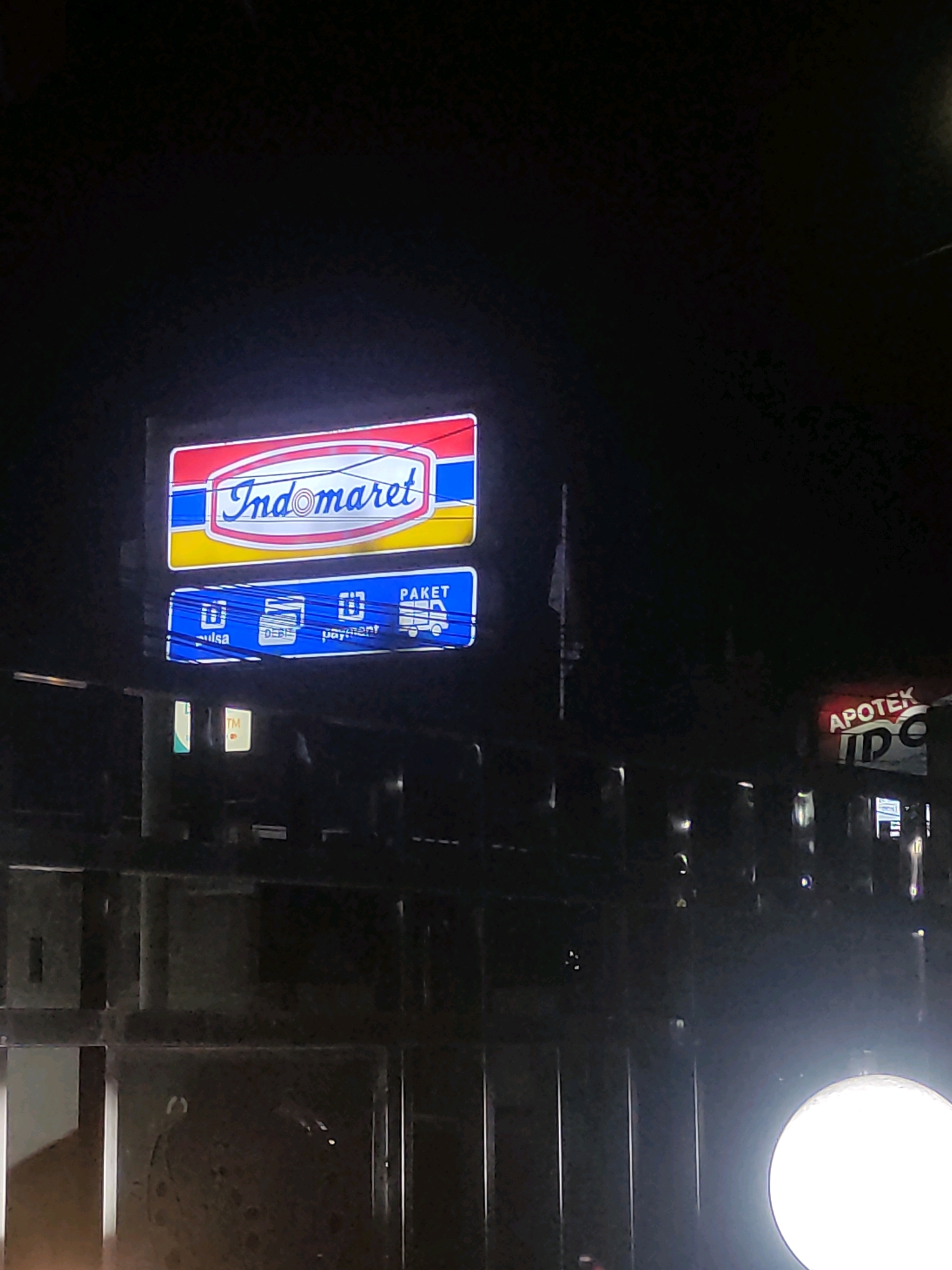 The image shows a brightly lit Indomaret sign at night in Ngasem, Indonesia. The sign is rectangular with a red border and features the Indomaret logo in blue and white. Below the main sign, there is a smaller blue sign with icons and text that appear to indicate services such as "pulsa" (credit), "debit," "payment," and "paket" (package delivery). To the right of the Indomaret sign, there is another illuminated sign that reads "APOTEK" (pharmacy) followed by some unreadable text. The signs are mounted on a dark background, likely a building, and are illuminated against the night sky. In the foreground, there is a dark, blurred structure that appears to be a fence or gate. The overall scene suggests a commercial area at night.