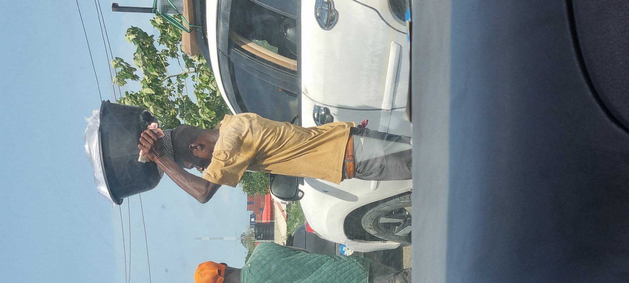 The image shows a man standing next to a white car, holding a black bucket filled with a plastic bag and some currency notes over his head in Satellite Town, Nigeria. He is wearing a yellow shirt and gray pants. Another person wearing a green jacket and orange cap is standing slightly behind him. There is a building and trees visible in the background.