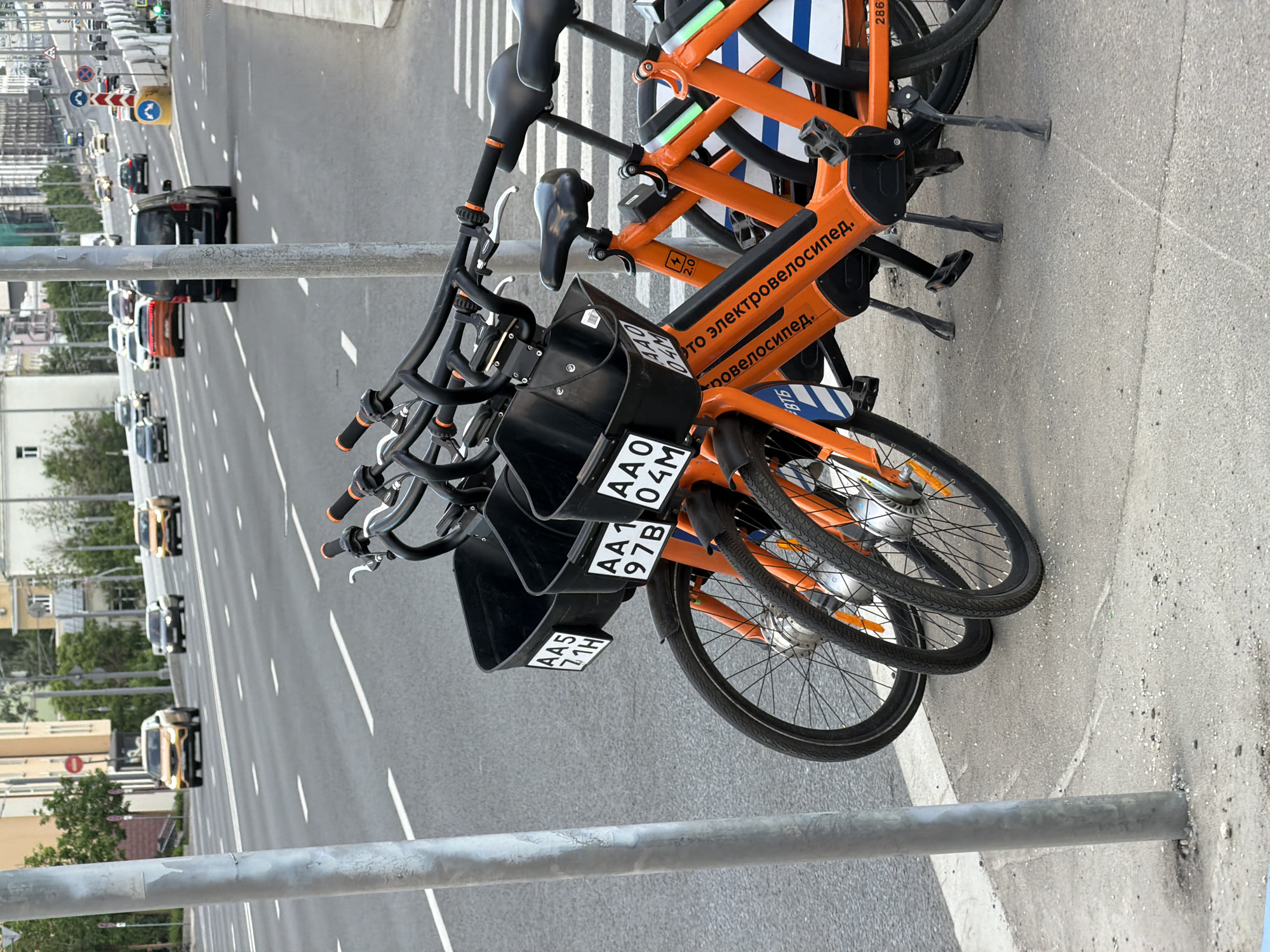 Here's a detailed description of the image:
**Scene Overview:**
The image captures a street scene in what appears to be an urban environment. The camera is positioned at street level, looking at a row of orange-colored rental bicycles. The bikes are parked on the edge of the sidewalk, with a busy road and buildings in the background.
**Subjects and Objects:**
* **Bicycles:** There are at least two rental bicycles in the foreground, primarily orange, with black handlebars and baskets. There are license plates on the front of the bikes with the numbers and letters: AA5 71H, AAO 97B, and AA1 04M. The text on the side of the bike reads "Это электровелосипед" (This is an electric bike) in Russian.
* **Road:** A multi-lane road is visible in the background, with vehicles traveling in both directions. There are lane markings and other road signs.
* **Sidewalk:** The bikes are parked on the edge of a concrete sidewalk.
* **Background:** Buildings and trees are visible in the distance.
**Environment and Location:**
The environment appears to be a bustling urban area. Based on the language and license plate on the bicycles, the location is likely in Russia.
**Additional Observations:**
* The lighting suggests it is daytime.
* The image is angled, giving a slightly tilted perspective.