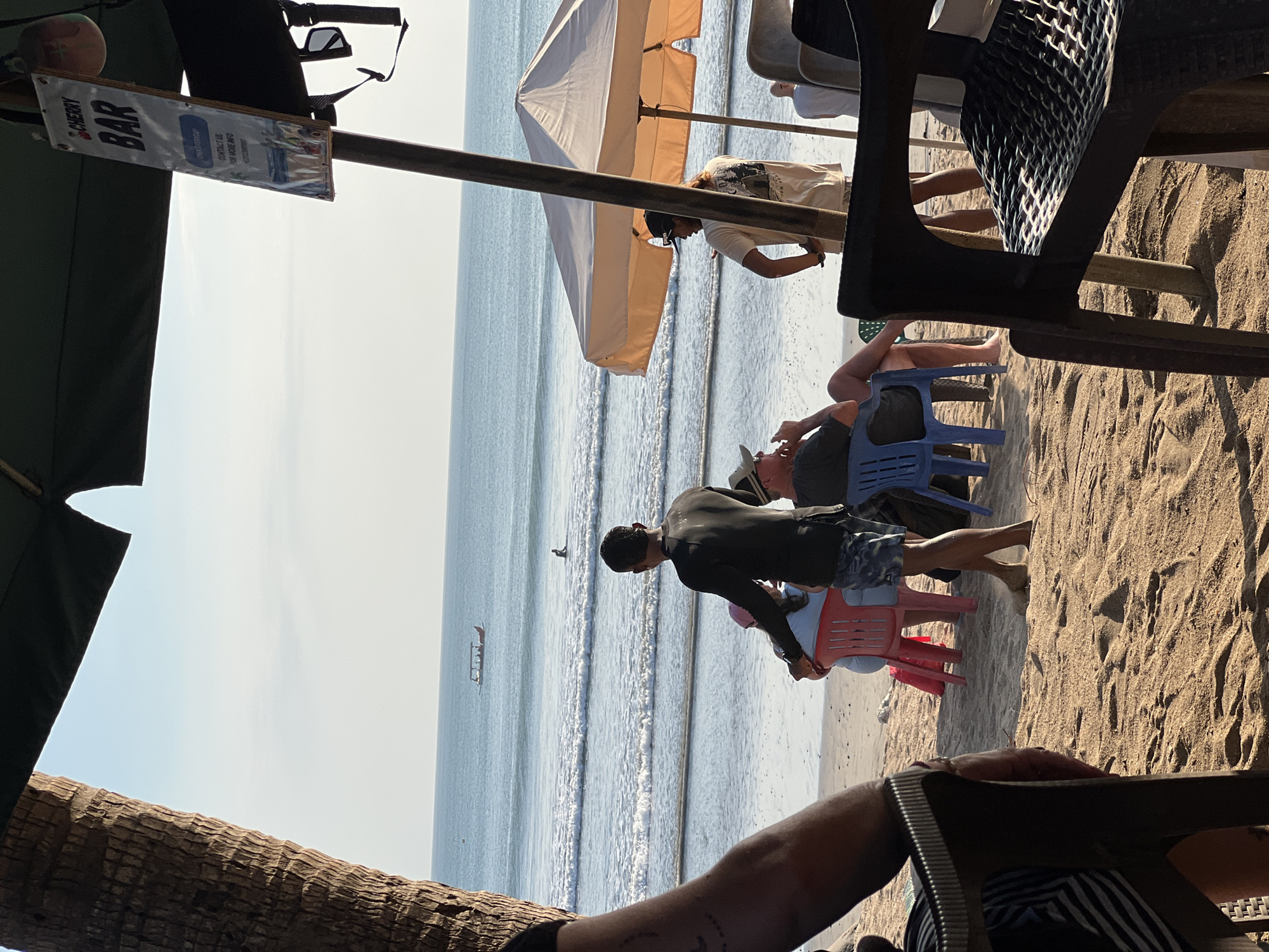 A beach bar scene is depicted in Kuta, Indonesia, on a clear day. In the foreground, a person's arm and shoulder are visible, seated in a chair. Behind them, several other individuals are seated or standing on the sandy beach. One man, wearing a dark long-sleeved shirt and patterned shorts, stands near some red chairs, appearing to interact with someone seated. Further back, a man in a light-colored t-shirt and shorts stands near a beige umbrella, looking towards the ocean. Another person in a white shirt is visible in the distance. The ocean is calm with small waves, and a longboat with passengers is visible on the water. A sign for "Cherry Bar" is hanging from a pole. The environment suggests a relaxed atmosphere, characteristic of a beachfront establishment.
