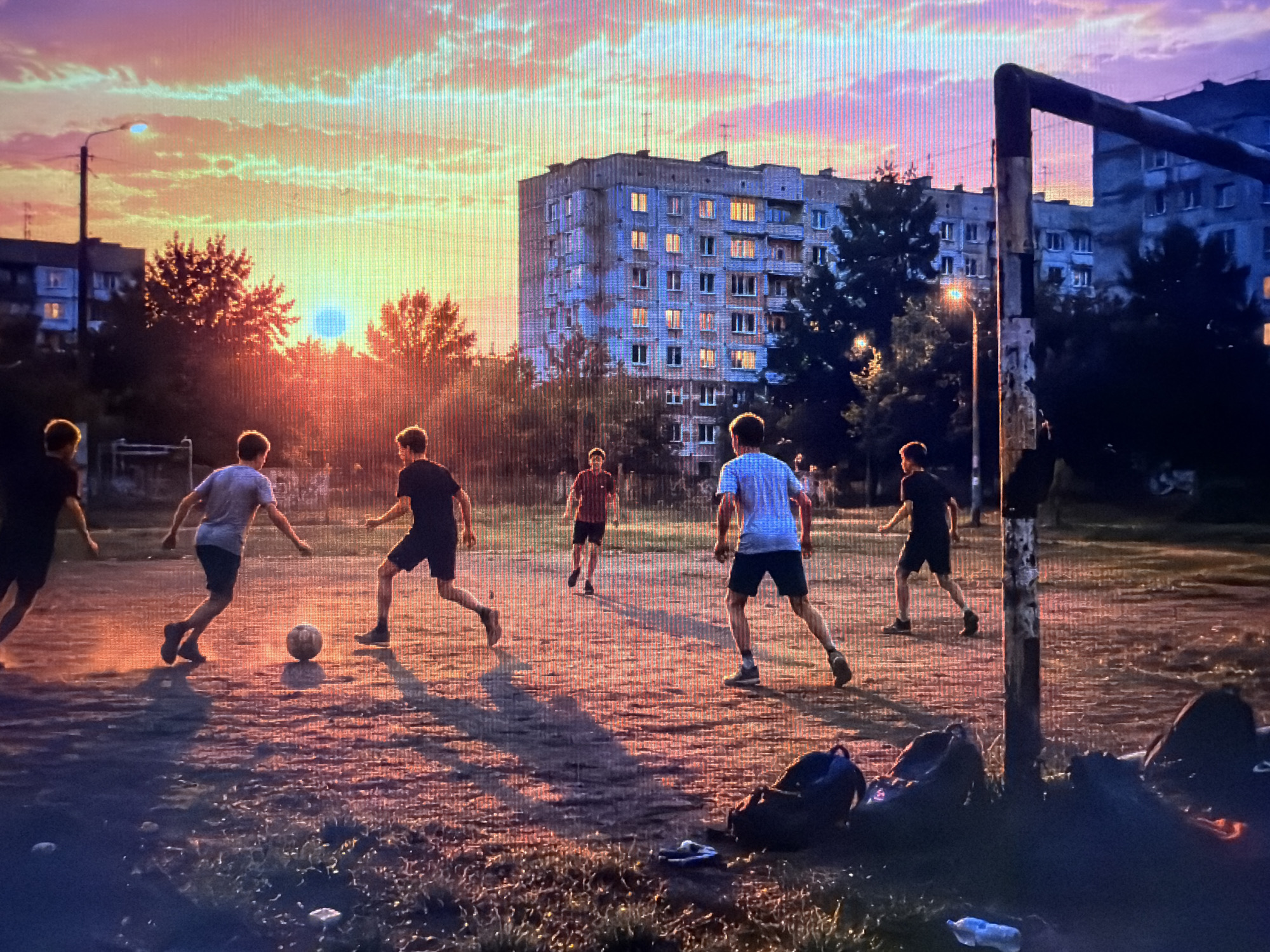This media file depicts a group of young people playing soccer on a dirt field at dusk. The sky is filled with vibrant colors of orange, pink, and purple, indicating either sunrise or sunset, with the sun appearing as a bright, hazy orb. Silhouetted against the colorful sky are apartment buildings, trees, and a streetlamp. The figures are engaged in an active game of soccer, with one person kicking the ball and others running or positioned to receive it.
Notable details include the rough texture of the playing field, the long shadows cast by the players due to the low angle of the sun, and the presence of several backpacks and a plastic bottle lying on the ground near a makeshift goalpost. The overall mood of the scene is one of youthful energy and casual recreation, set against a picturesque, urban twilight backdrop. The location appears to be a residential area with older, utilitarian apartment blocks. No visible text is present in the image.