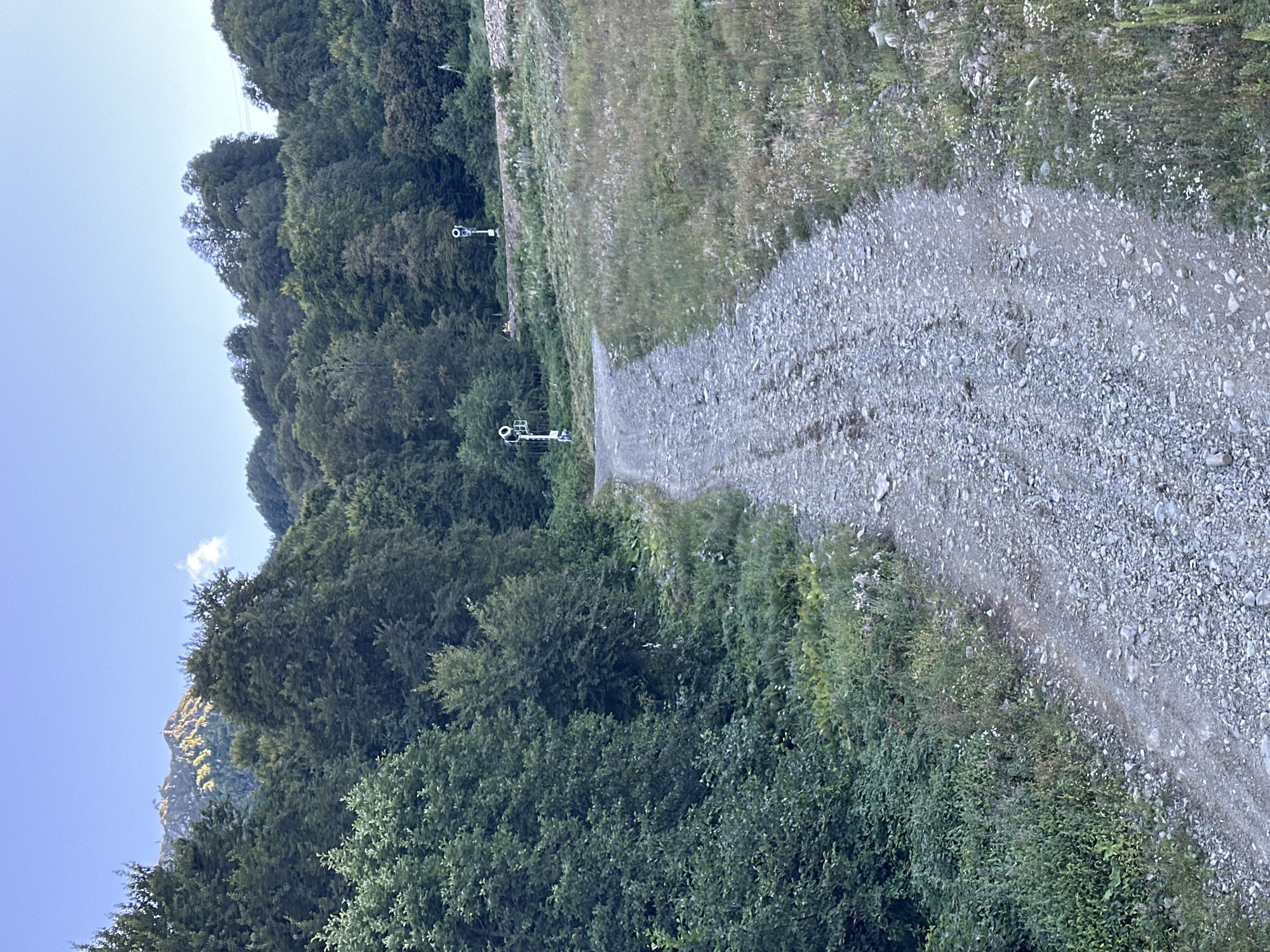 A dirt road winds uphill through a green mountain landscape under a clear blue sky. The road is covered in gravel and shows tire tracks. On either side of the road are dense green trees and vegetation. In the distance, at the top of the hill, are two sets of railway signals. The mountain range in the far background is illuminated by sunlight, with a few clouds in the sky. The scene is likely located in or around Estosadok, Russia, a mountainous resort town. There are no people visible in the image.