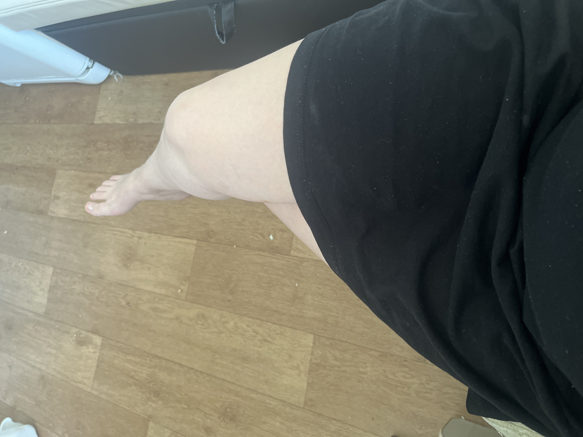 The image shows a person's legs and feet, viewed from above. The person is wearing a black skirt or dress. One leg is extended, with the foot visible. The other leg is bent. The person is sitting or lying on a wooden floor, which appears to be laminate flooring. There is a white object, possibly a piece of furniture or a blanket, in the upper right corner of the image. The overall scene suggests an indoor domestic setting. No specific details about the city of Chelyabinsk, Russia, can be inferred from the image.