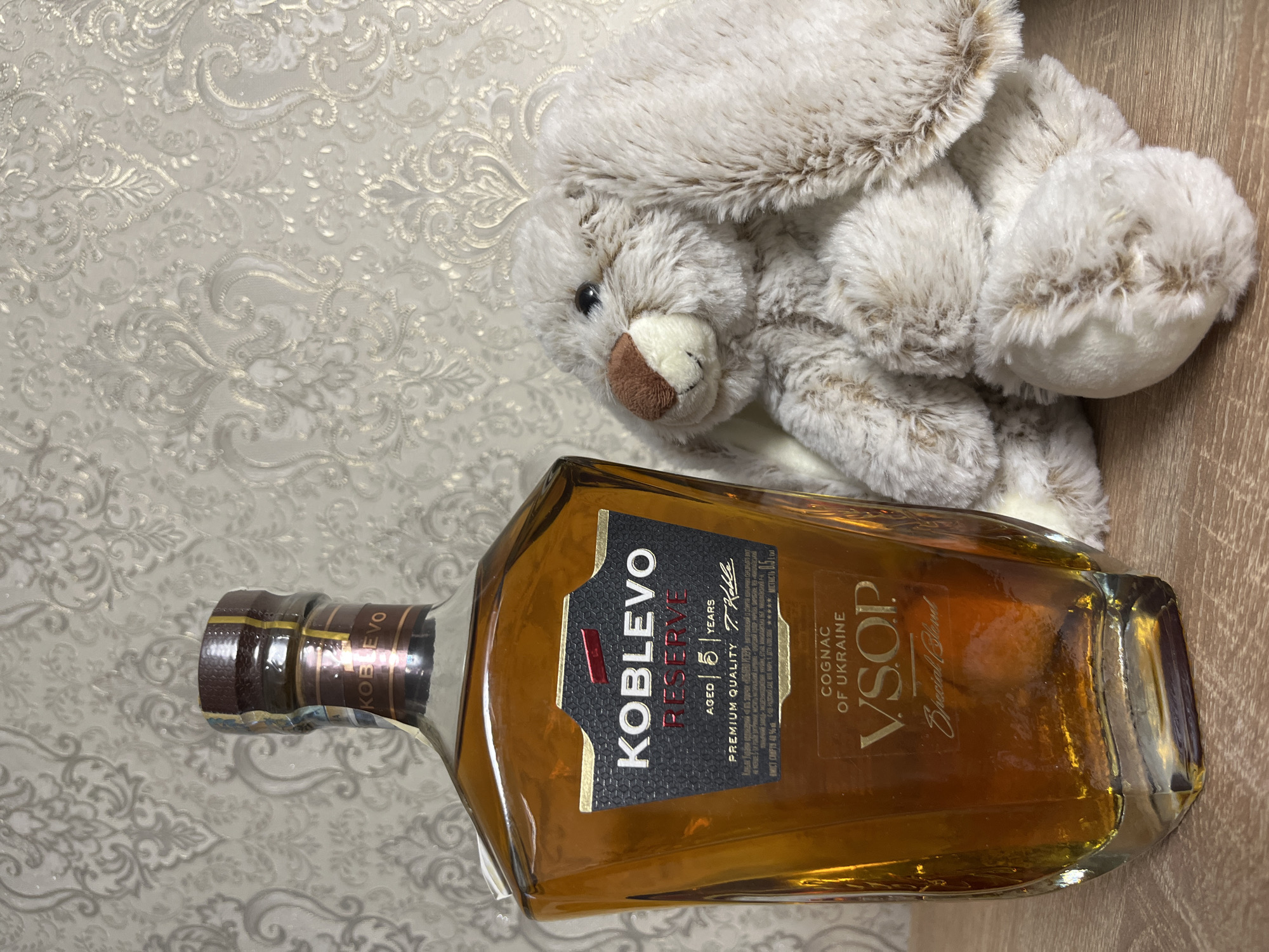 An indoor shot taken in Kryvyi Rih, Ukraine, features a bottle of "KOBLEVO RESERVE" cognac and a light-colored plush bunny toy. The amber-hued bottle displays a black and gold label with text including "KOBLEVO RESERVE," "AGED 5 YEARS," "PREMIUM QUALITY," "COGNAC OF UKRAINE," and "V.S.O.P. Special Blend." Further details on the label indicate "MICT COMPTS 40%" (likely alcohol percentage) and "MІСТКІСТЬ 0.5" (likely volume, 0.5 liters). The bottle's cap is a metallic brown with gold accents. The plush bunny, predominantly white with light brown tips on its fur, is lying next to the bottle. The background consists of a light-toned wallpaper with an intricate, shimmering, silver-grey floral and swirl pattern. A plain light brown wooden surface is visible beneath the objects.