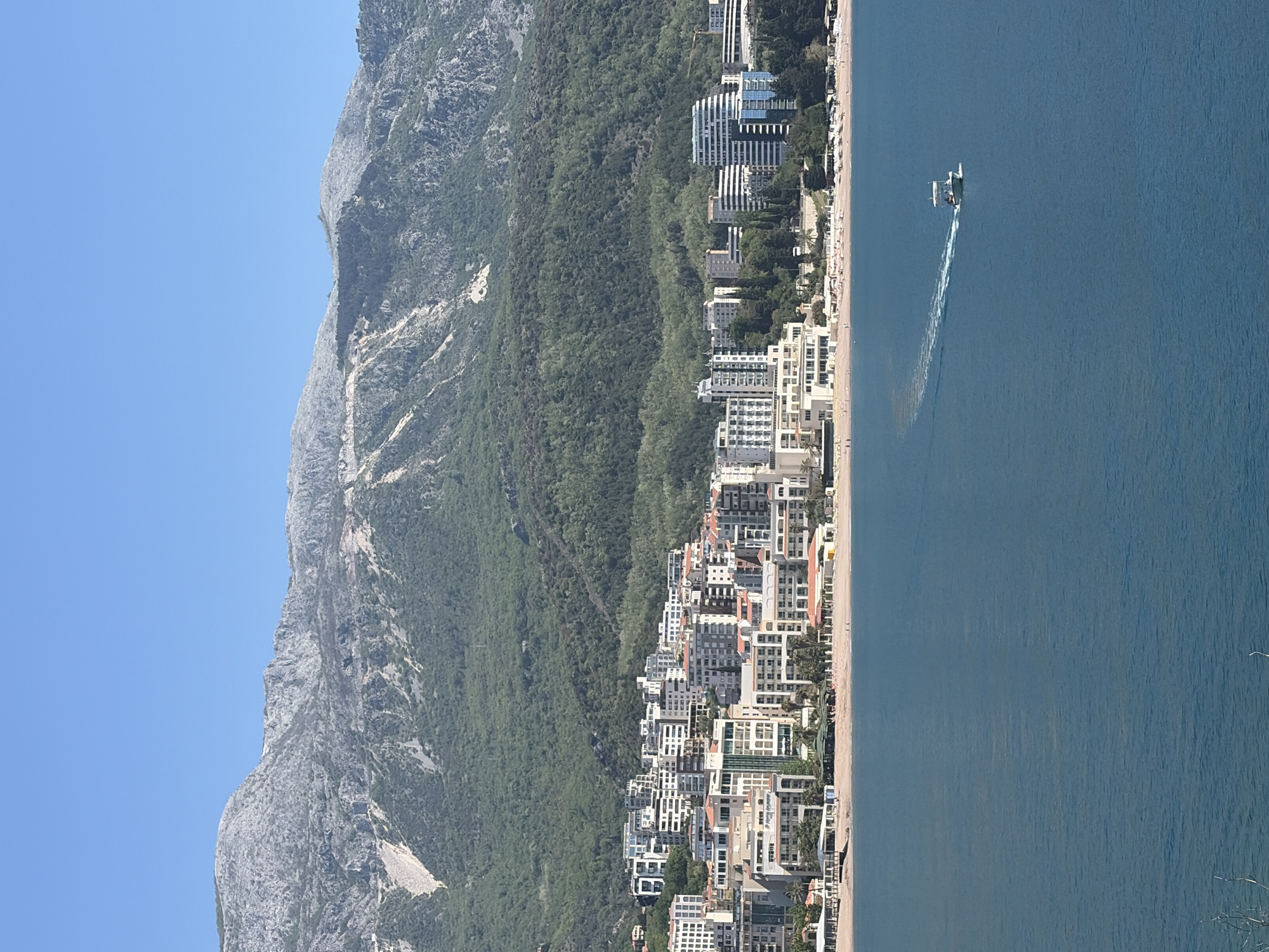 The image shows a coastal landscape in Budva, Montenegro. The Adriatic Sea occupies the right half of the image. A speedboat is visible on the water, leaving a trail. Along the coastline, there is a beach with buildings lining the shore. The buildings are a mix of residential and commercial structures. A mountain covered in green vegetation rises up on the left side of the image. The sky is clear and blue.