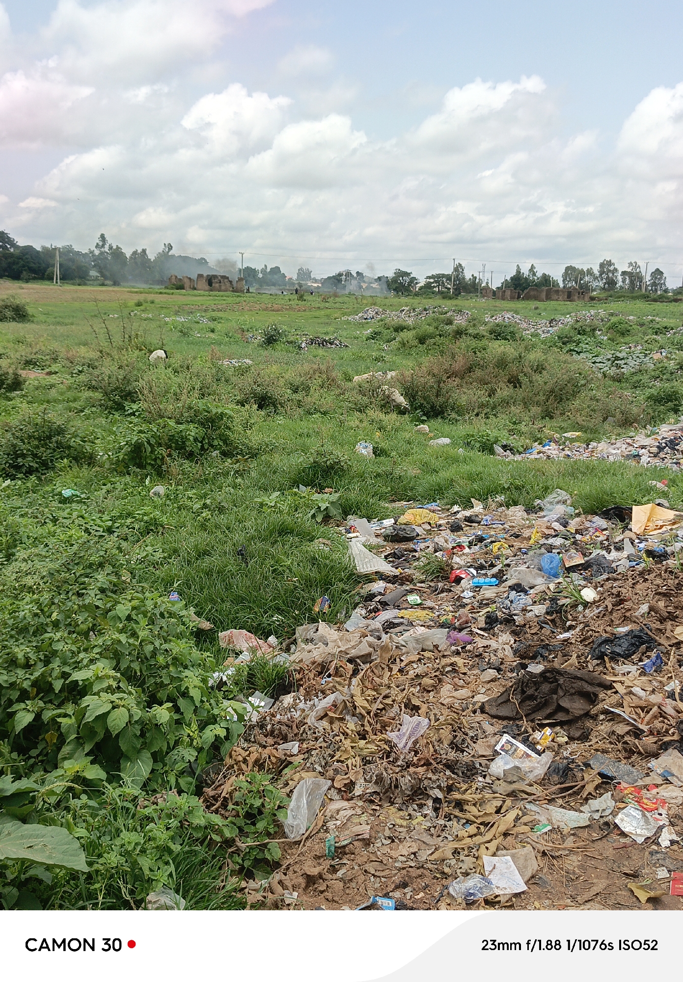 A wide-angle photograph captures a desolate landscape filled with discarded trash and overgrown vegetation under a cloudy sky. In the foreground, a large pile of garbage dominates the right side of the frame, consisting of plastic bags, paper waste, food scraps, and other refuse. The debris is a mix of colors, with browns, grays, and some splashes of red and blue from plastic items. To the left of the garbage pile, lush green plants and grass are visible, growing in abundance.
In the middle ground, the landscape transitions to a grassy field with scattered bushes and more piles of trash in the distance. Several dilapidated structures, possibly abandoned buildings or ruins, are visible in the far background, with smoke rising from them, suggesting a potential fire or industrial activity. A few sparse trees line the horizon. The sky is overcast with large, puffy white clouds. No people are clearly visible in the scene. The overall impression is one of neglect and environmental degradation in a rural or semi-urban setting.