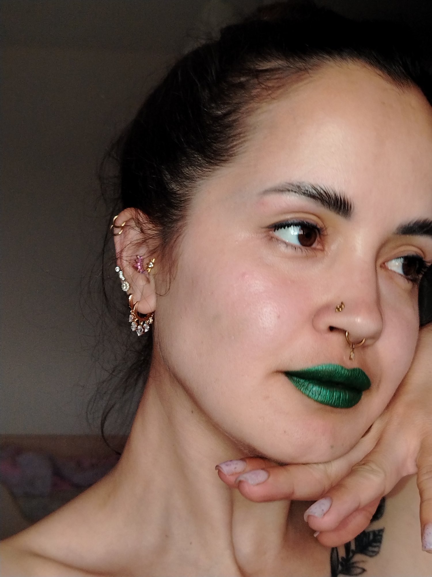 A close-up indoor portrait captures a woman from her forehead to her upper chest. She has dark hair, likely pulled back, and her dark eyes are directed towards the right side of the frame, suggesting a thoughtful or observant gaze. Her expression is neutral and serene.
A prominent feature is her metallic green lipstick. Her face is adorned with multiple piercings: a gold-toned stud on her right nostril and a gold septum ring with a small dangling charm. Her left ear is extensively decorated with various gold-toned piercings, including multiple small hoops along the upper cartilage, an ear climber with clear and pink stones, and a larger hoop earring in the lobe featuring several dangling clear stones.
Her left hand, with light-colored, somewhat chipped nail polish, rests gently against her chin and jawline. A portion of a dark tattoo, depicting what appears to be foliage or leaves, is visible on her lower neck or upper chest. The background is uniformly dark and out of focus, indicating an indoor setting, possibly in Noviy, Russia, with soft, directional lighting highlighting her features and jewelry. No text is visible in the image.