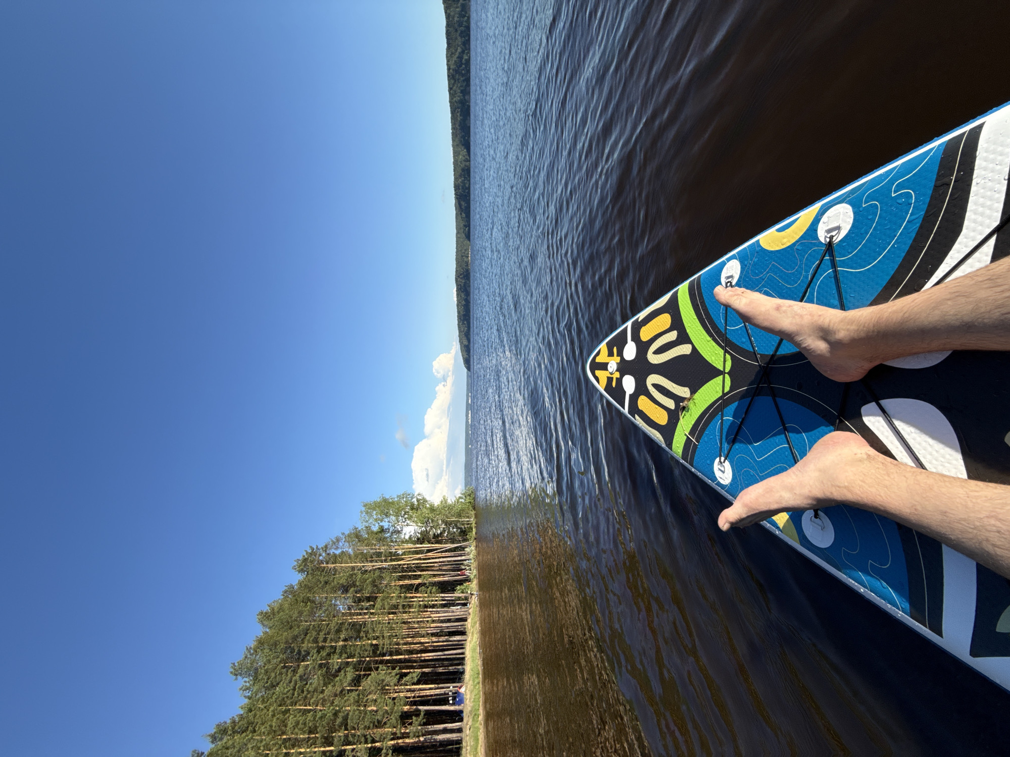 The image is a first-person perspective from a paddleboard on a body of water, likely a lake or river, near Gora (Khokhlovskoe S/P), Russia. The foreground shows the front of a paddleboard with a blue, white, yellow, and green design, and a person's bare feet positioned on it. The water is dark and choppy, with ripples spreading out from the board.
In the middle ground, the water extends to a tree-lined shore. The trees are tall and slender, predominantly pines, with some lighter foliage mixed in. The shore appears to be sandy or grassy, with a few people visible in the distance, engaged in recreational activities. Two blue tents are also present on the shoreline.
The background features a wide expanse of water meeting the horizon. On the far side of the water, another tree-covered bank or island is visible. The sky is a clear, bright blue with a few white clouds towards the horizon. The overall scene suggests a sunny day spent outdoors enjoying water activities.