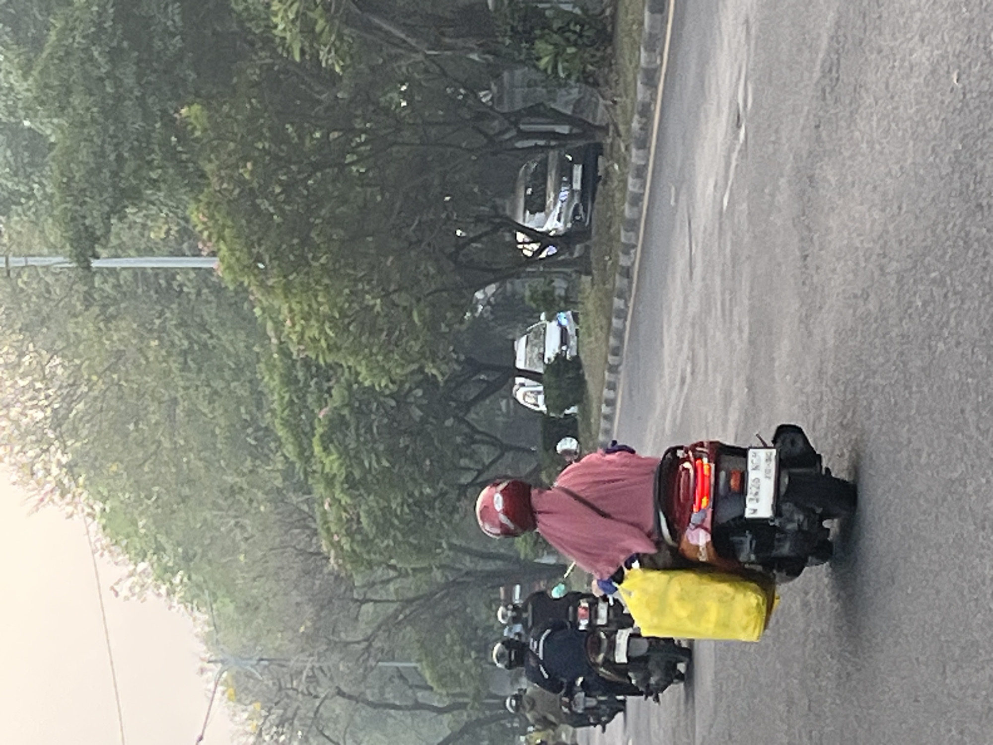 In Gelam, Indonesia, a group of people are seen riding motorbikes on a paved road lined with trees and a sidewalk. The foremost motorbike is ridden by an individual wearing a red helmet and a pink covering, carrying a bright yellow bag on the back. Several other motorbikes, each with one or two riders wearing helmets, are traveling ahead and behind. Two cars are visible in the background, parked on the side of the road. The lighting suggests it is daytime, with overcast conditions.