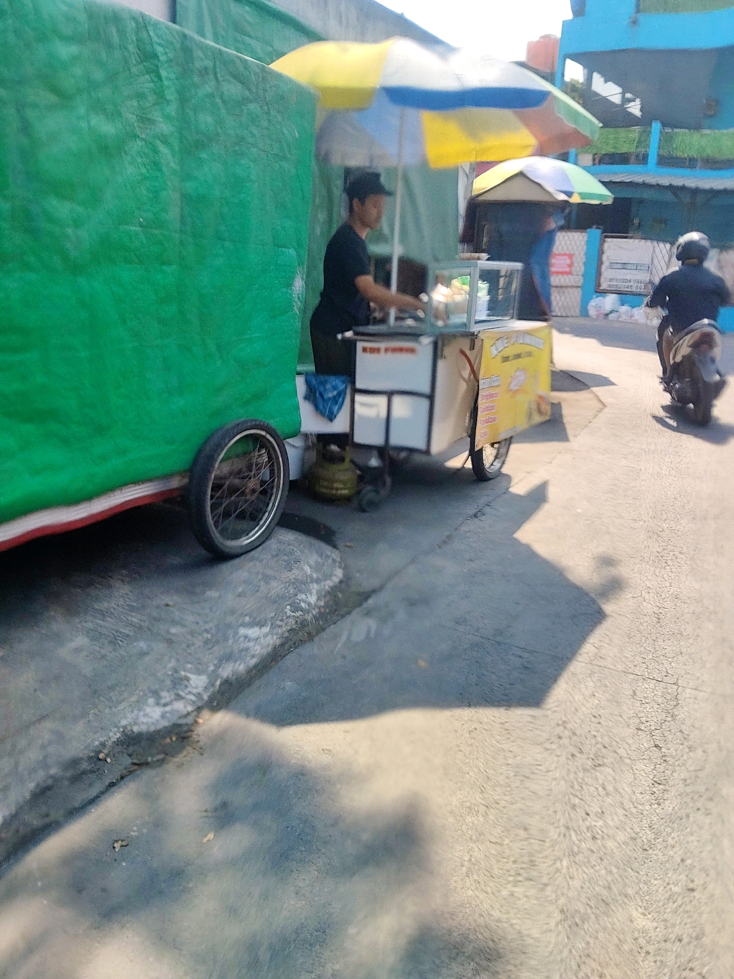 This is a low-angle, outdoor, eye-level photograph taken on a street in Mekarmukti, Indonesia. The scene depicts a food cart with a green tarp covering its left side. A person wearing a black shirt and dark pants is standing behind the food cart, preparing food. The cart has a glass display case and a yellow side panel with some text. A gas cylinder is visible on the ground next to the cart. A person is riding a motorcycle in the background, wearing a dark helmet and jacket. The street is paved, and shadows from an umbrella and possibly the food cart are cast on the ground. A building with blue accents is visible in the background, and a multi-colored umbrella is partially visible over the food cart. The overall lighting suggests it is daytime.