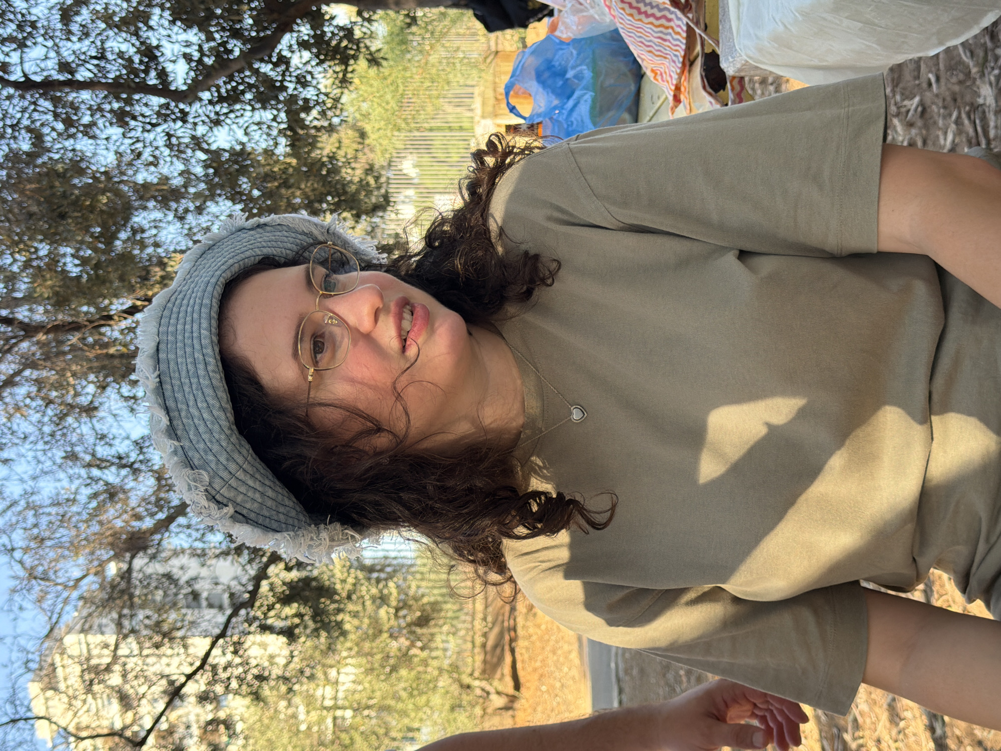 A young woman with curly brown hair and glasses is sitting outdoors, possibly at a picnic. She is wearing a light blue frayed bucket hat and a plain olive green t-shirt. A silver heart-shaped pendant necklace is visible around her neck. The background shows trees, a building, and a blue plastic bag on a table. The lighting suggests it is late afternoon, with shadows cast across the woman. The location is identified as Rishon LeZion, Israel.